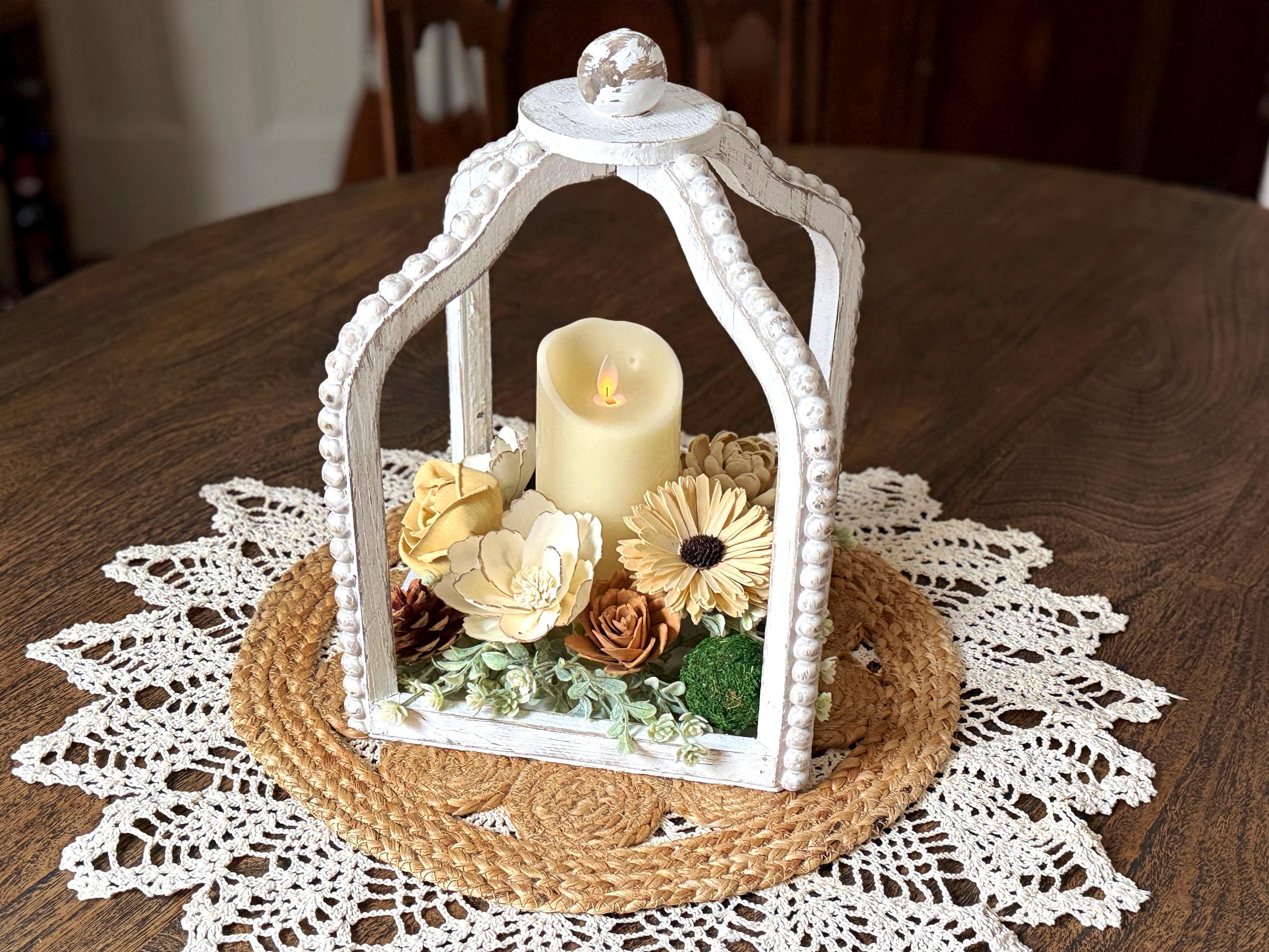 Decorative white lantern with a candle and flowers on a wooden table with a doily.