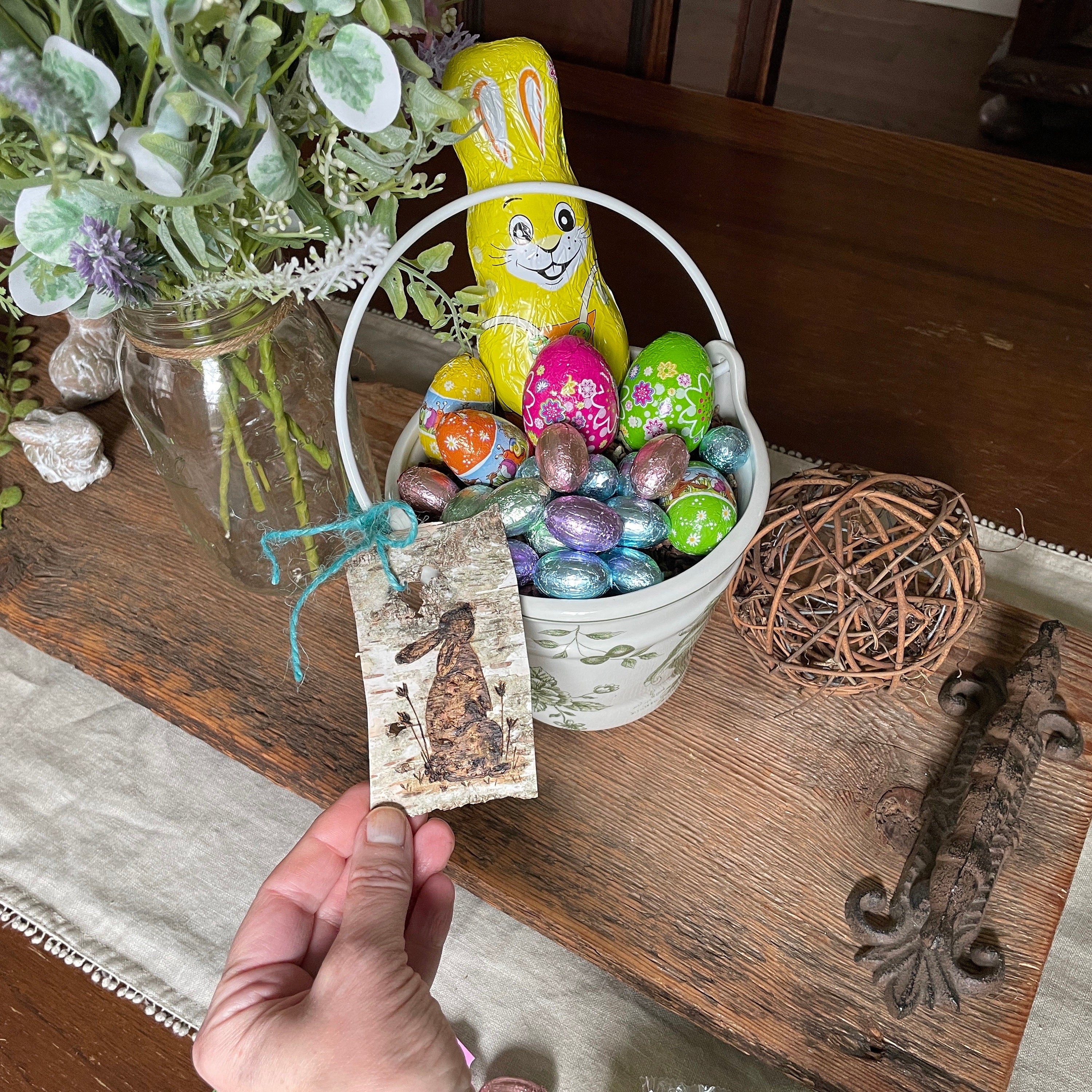 Decorative Easter basket with colorful eggs, a bunny tag, and a hand holding a tag on a wooden surface.