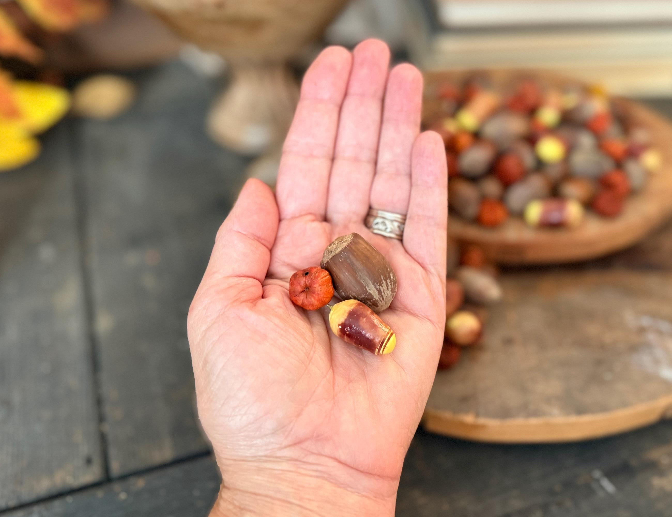 Hand holding three small, colorful objects included with bowl filler mix. Includes a pumpkin pod, a hazelnut and a faux candy corn. A blurred background of more similar items.