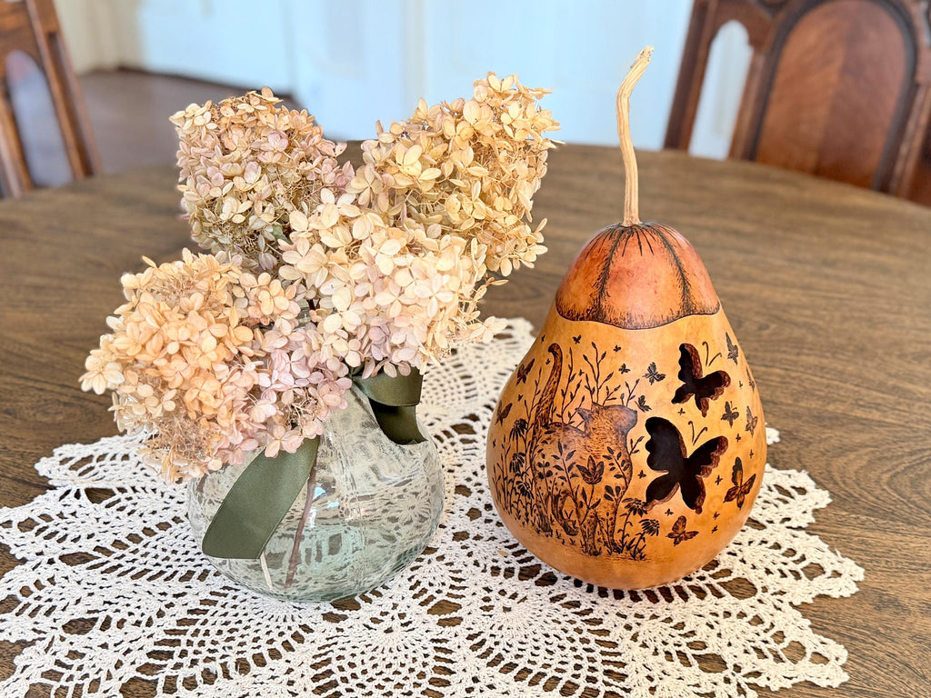 Decorative gourd with butterfly designs on a table with flowers and a doily.