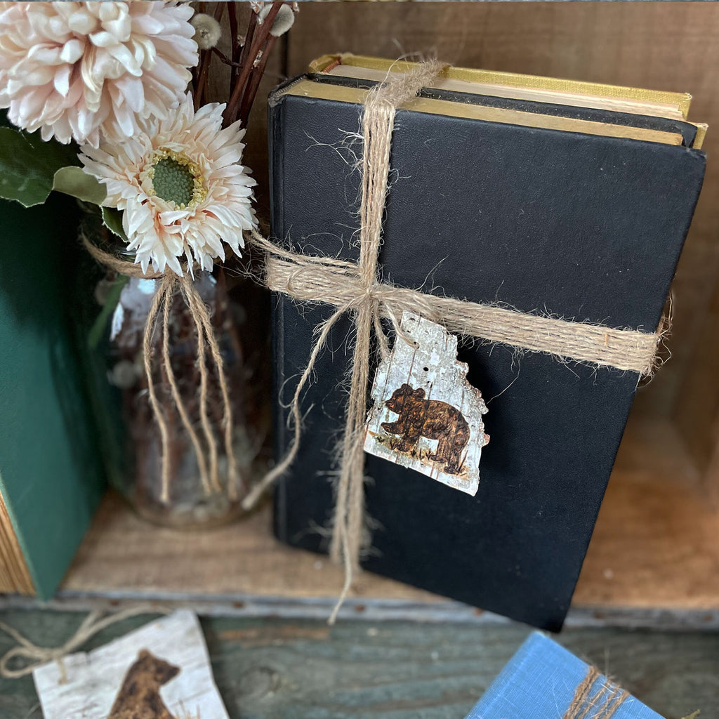 Black book tied with twine featuring a bear accent tag, placed on a wooden surface with flowers and books.