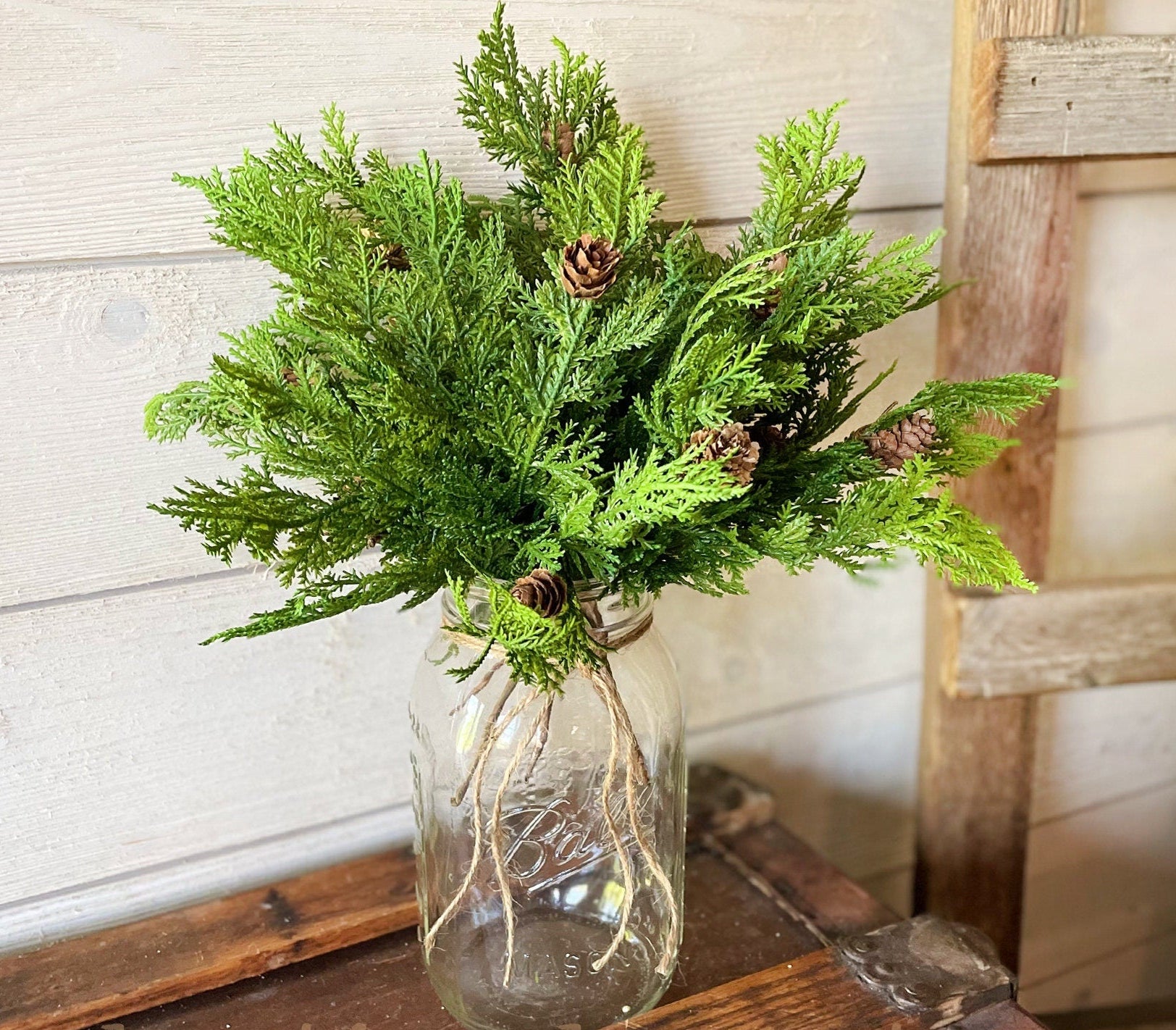 Bouquet of greenery in a clear glass jar on a wooden surface with a rustic background.