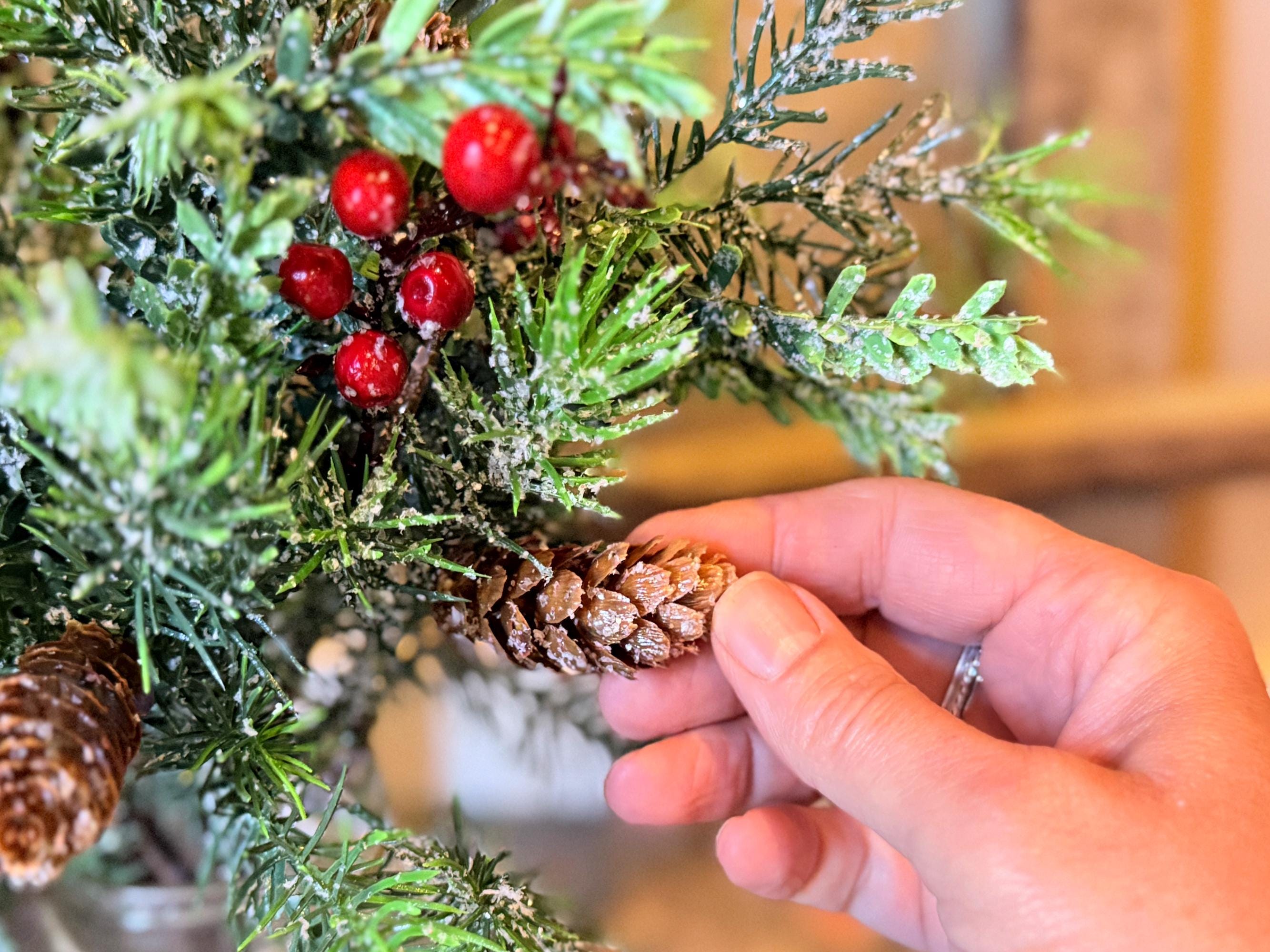 Hand holding a pine cone next to a festive wreath with greenery and red berries.