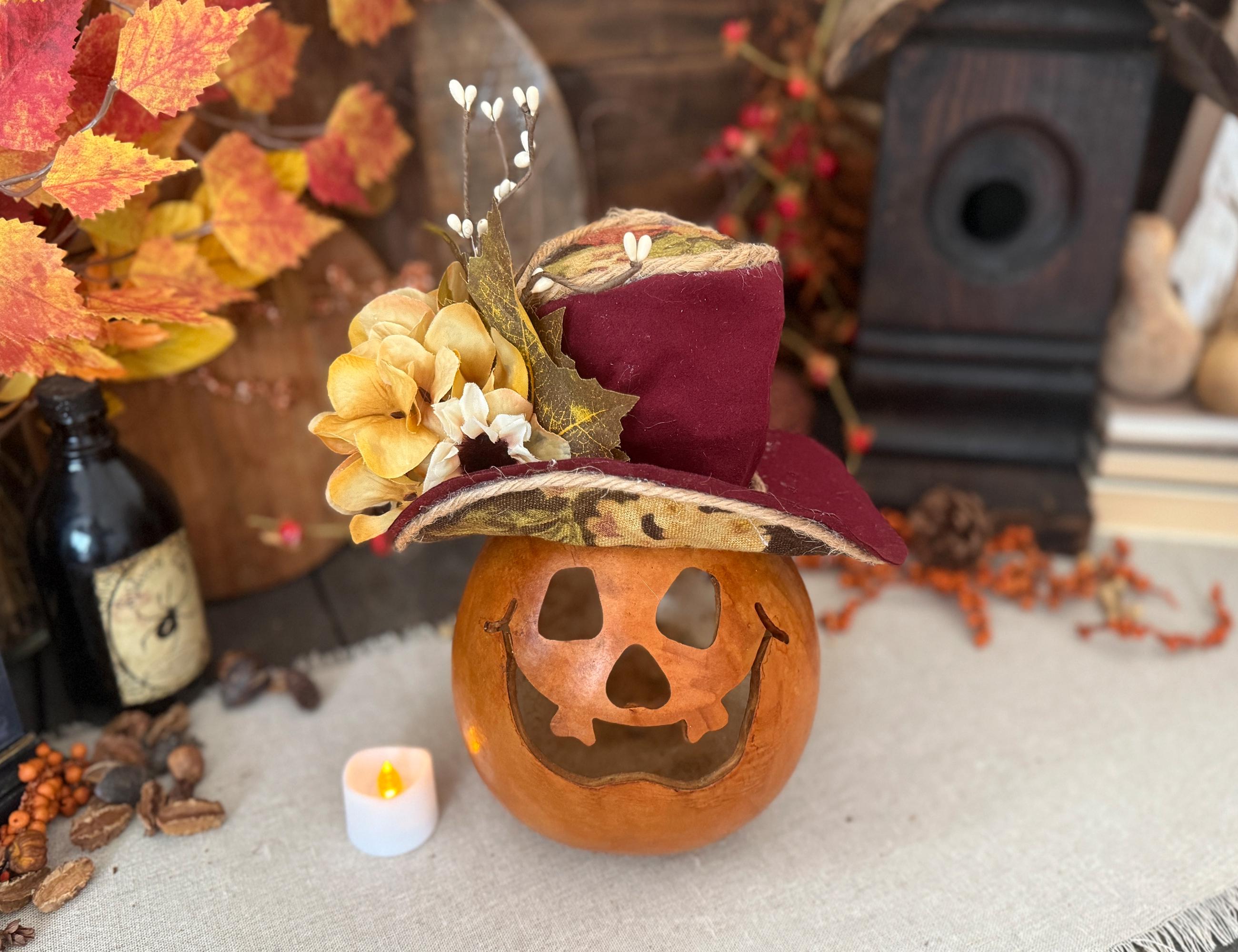 Decorative pumpkin with a carved face and top hat on a surface with autumn leaves and candles.