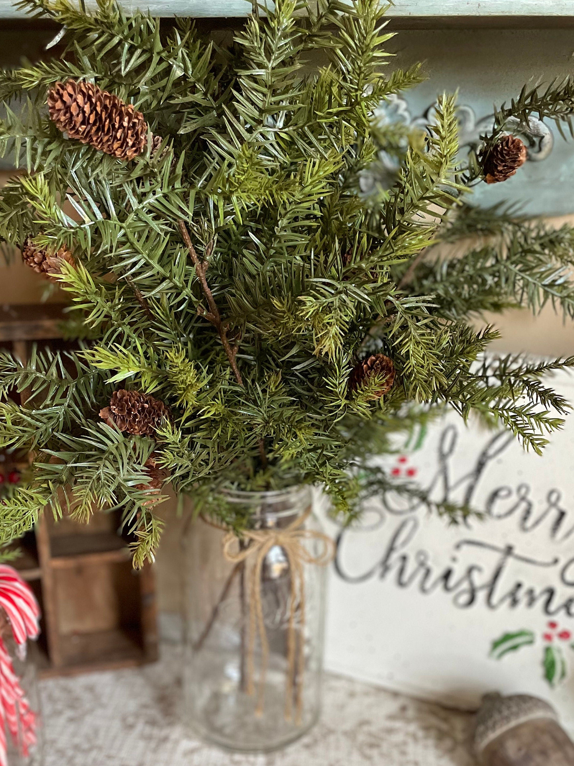 Decorative Christmas arrangement with greenery and pinecones in a jar, with a 'Merry Christmas' sign in the background.