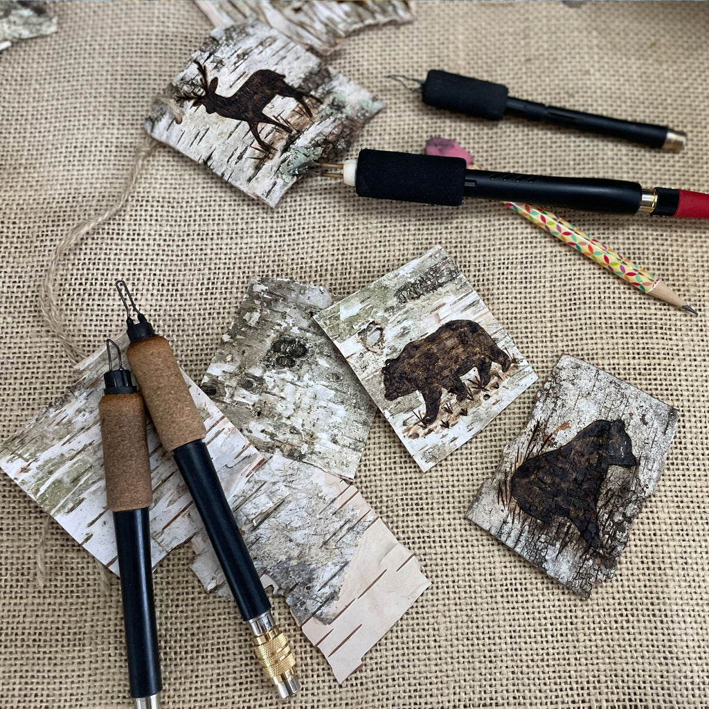 Artistic present toppers with animal designs on white birch bark, surrounded by carving tools on a textured fabric background.