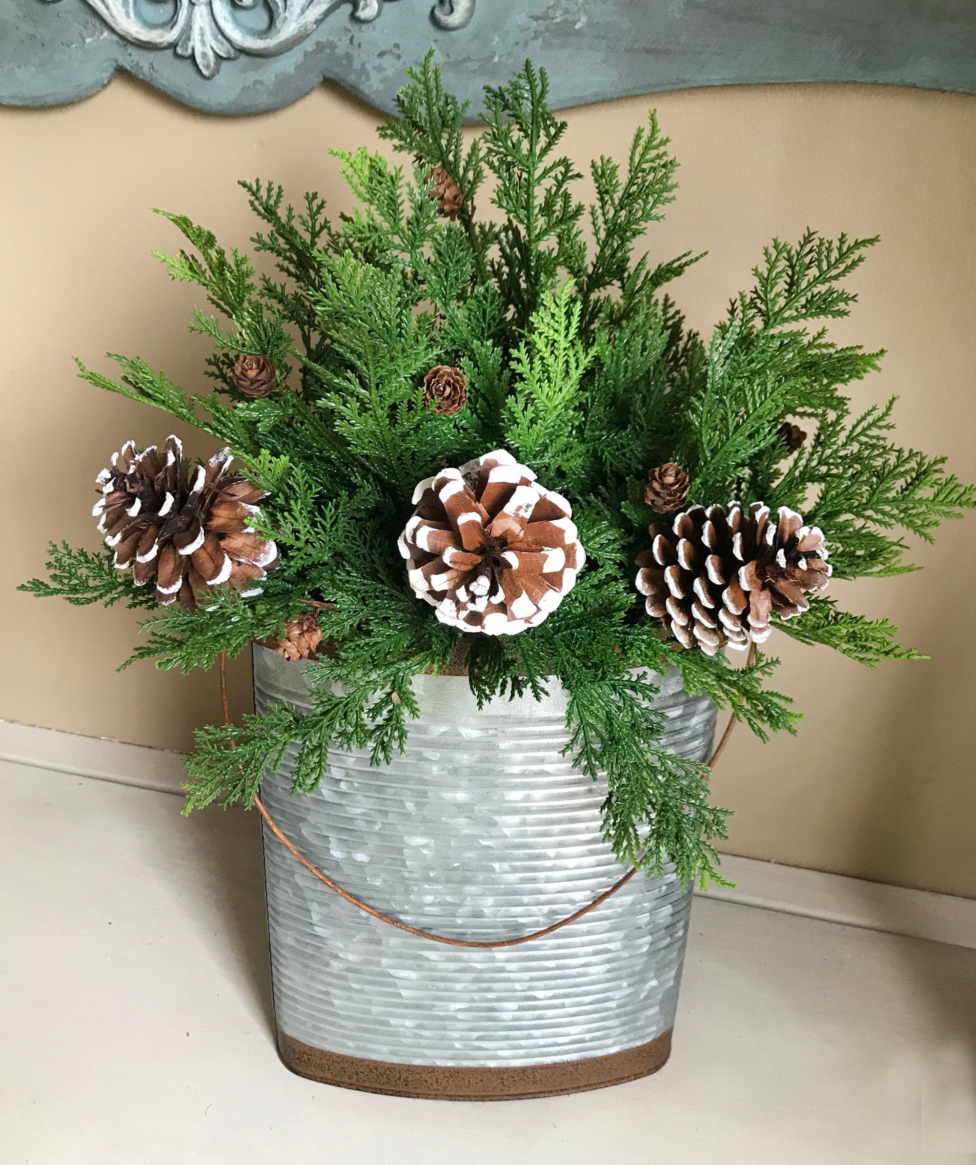 Decorative arrangement of greenery and pinecones in a metal container on a neutral background