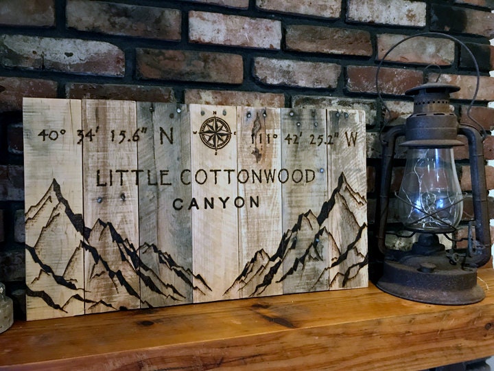 Wooden sign with mountain design and text 'Little Cottonwood Canyon' on a brick wall background.