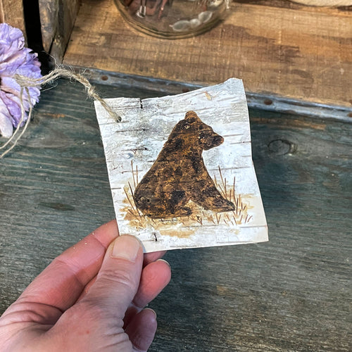 Hand holding a square birch bark ornament with a bear design on a rustic wooden surface.