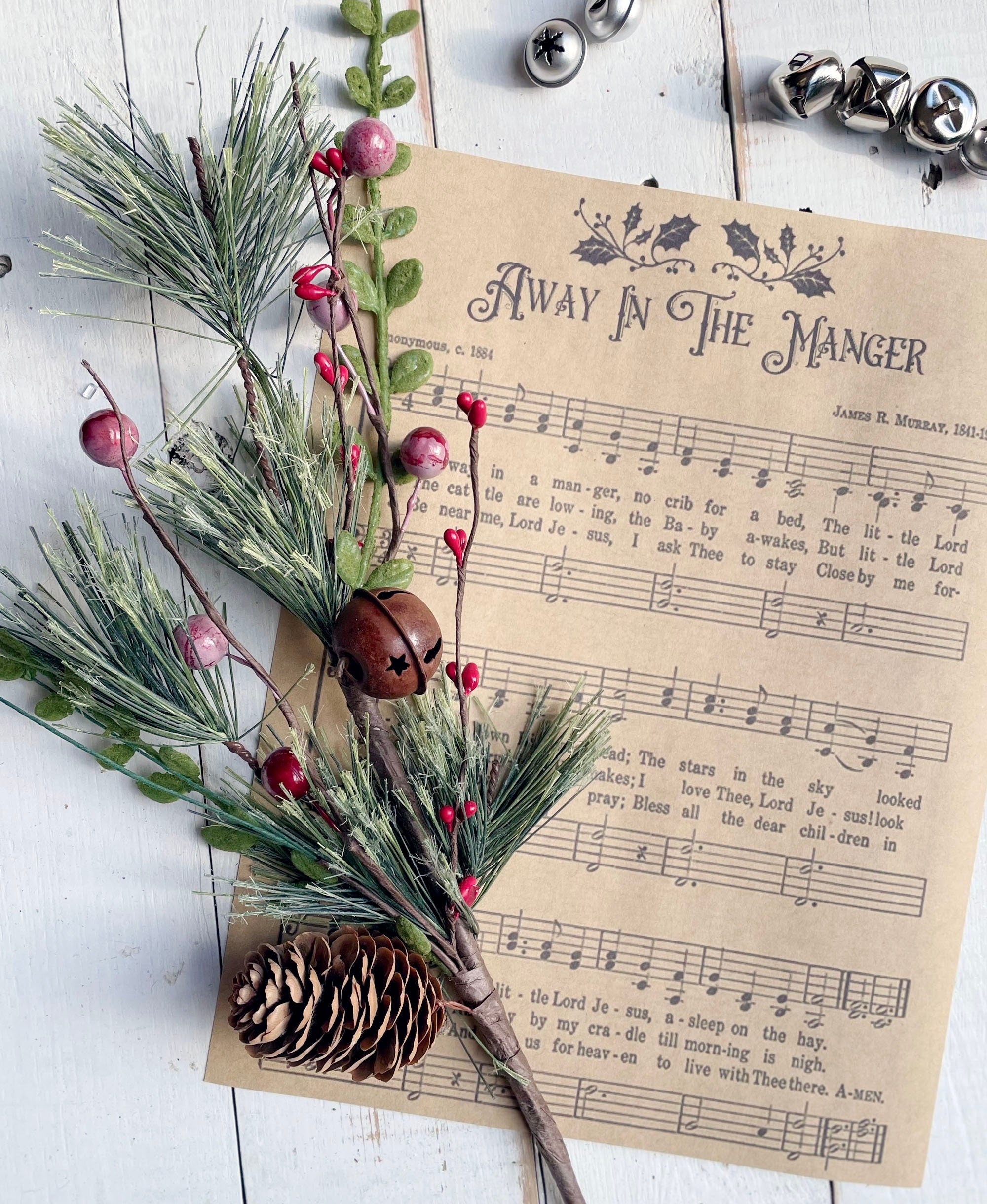 Decorative arrangement with greenery, berries, and pine cones on a sheet of music with 'Away in the Manger' text.