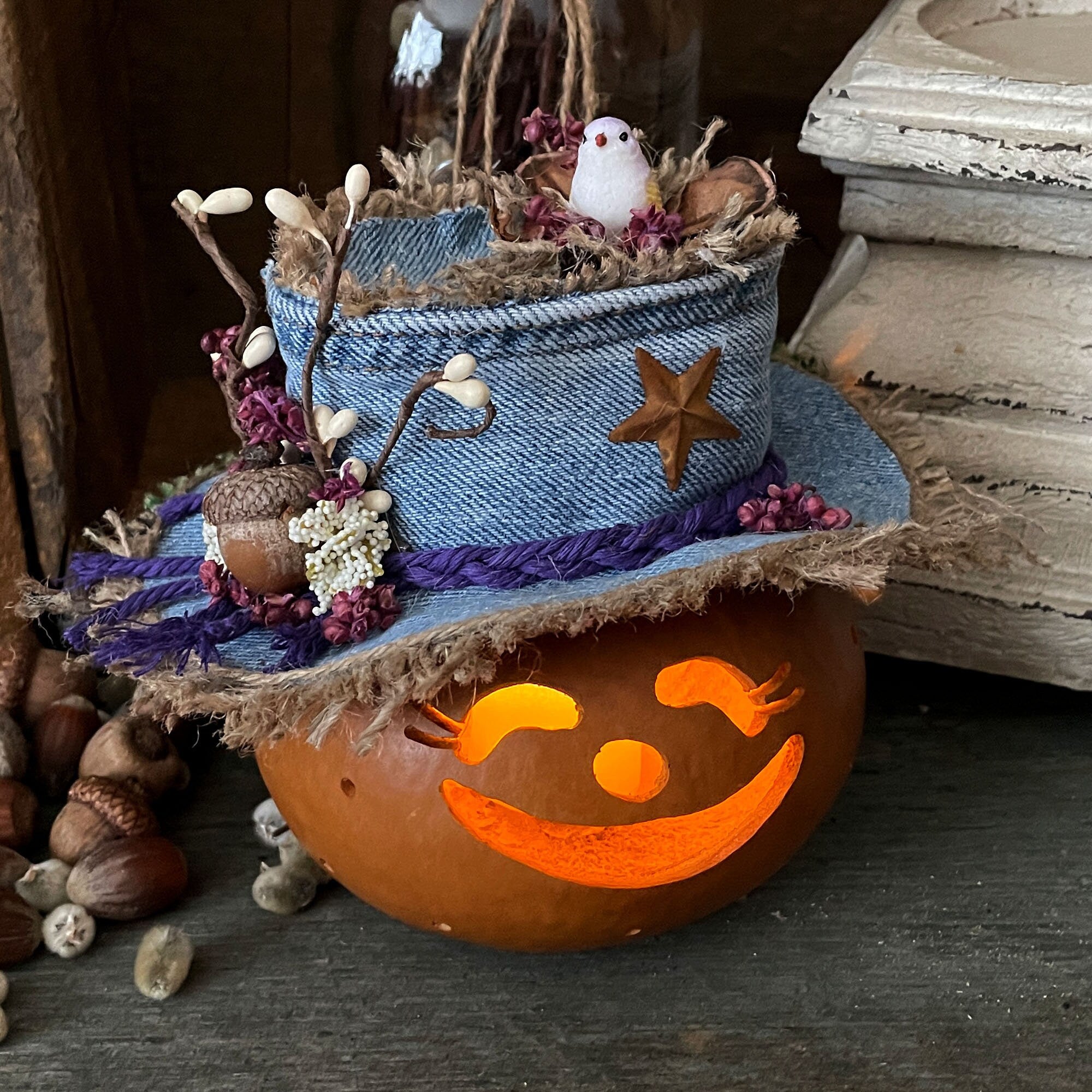 Decorative pumpkin with a face, surrounded by small plants and a blue woven basket on a wooden surface.