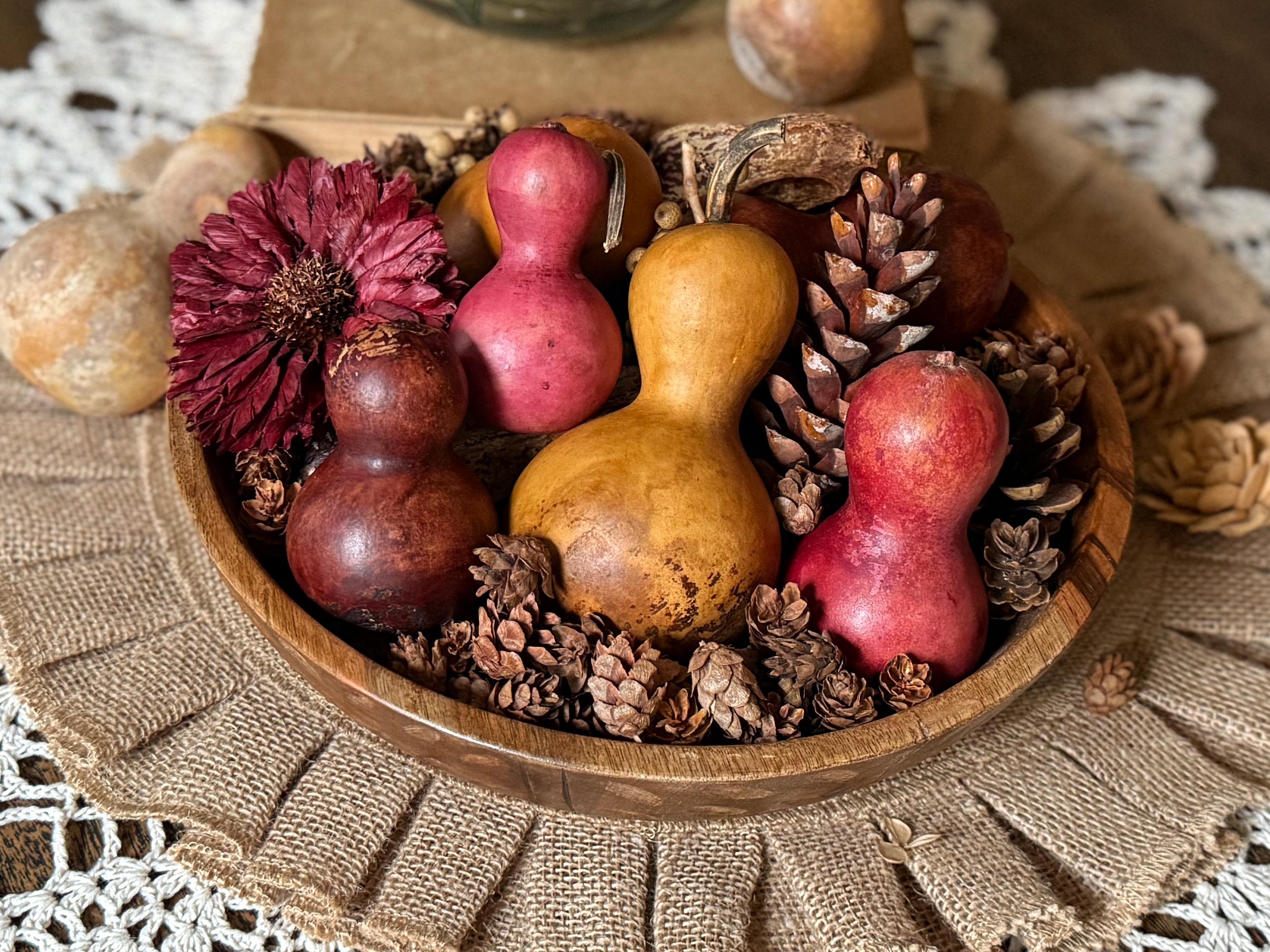 Decorative arrangement of gourds, pinecones, and flowers in a wooden bowl on a textured surface.