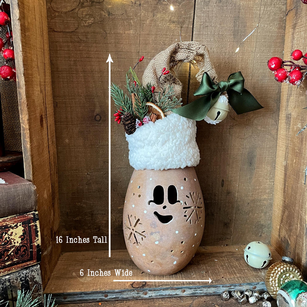 Decorative lantern with a face, Santa hat, and greenery on a wooden surface.