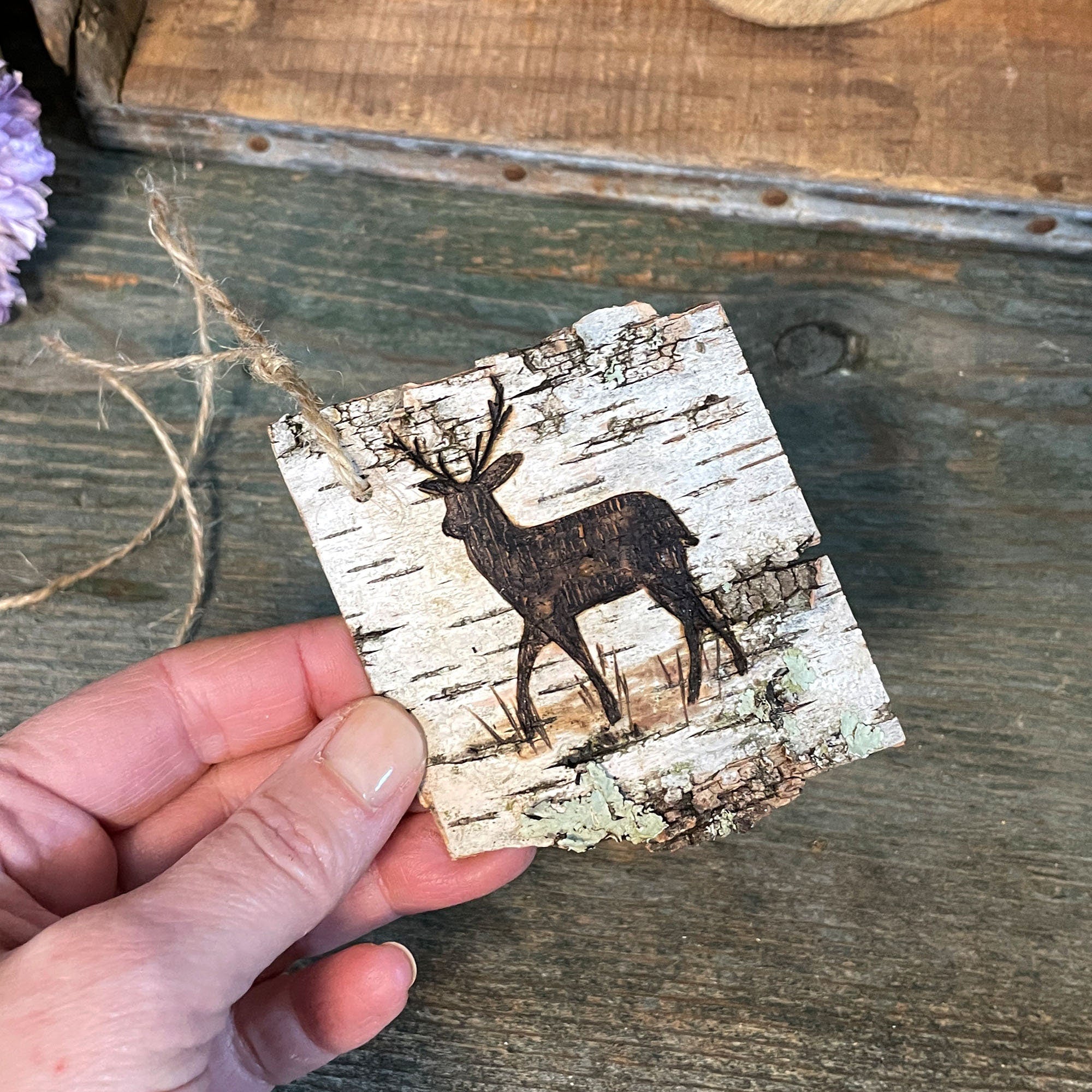 Hand holding a square birch bark ornament with a deer design on a rustic wooden surface.