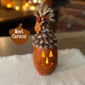 Decorative pumpkin with pine cone roof and glowing interior on a soft surface.