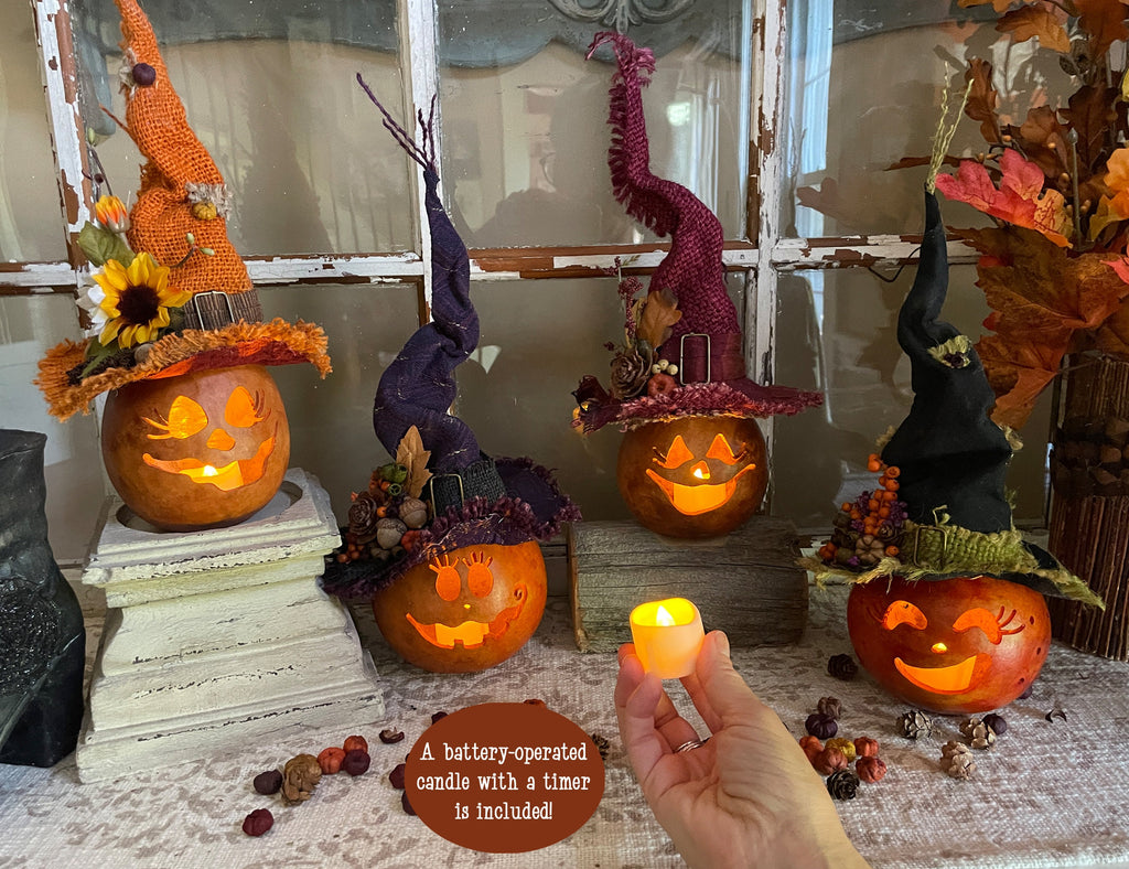 Decorative pumpkins with witch hats on a table, with a hand holding a small candle.