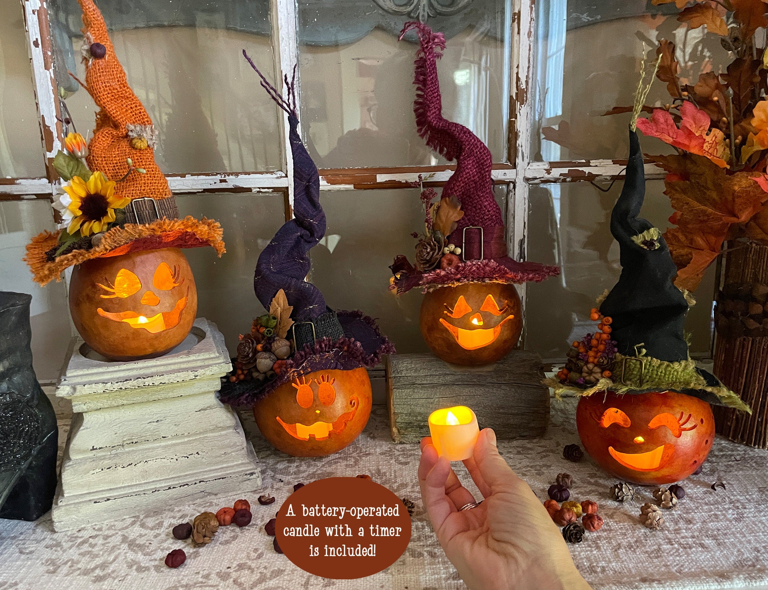 Decorative pumpkins with witch hats on a table, with a hand holding a small candle.