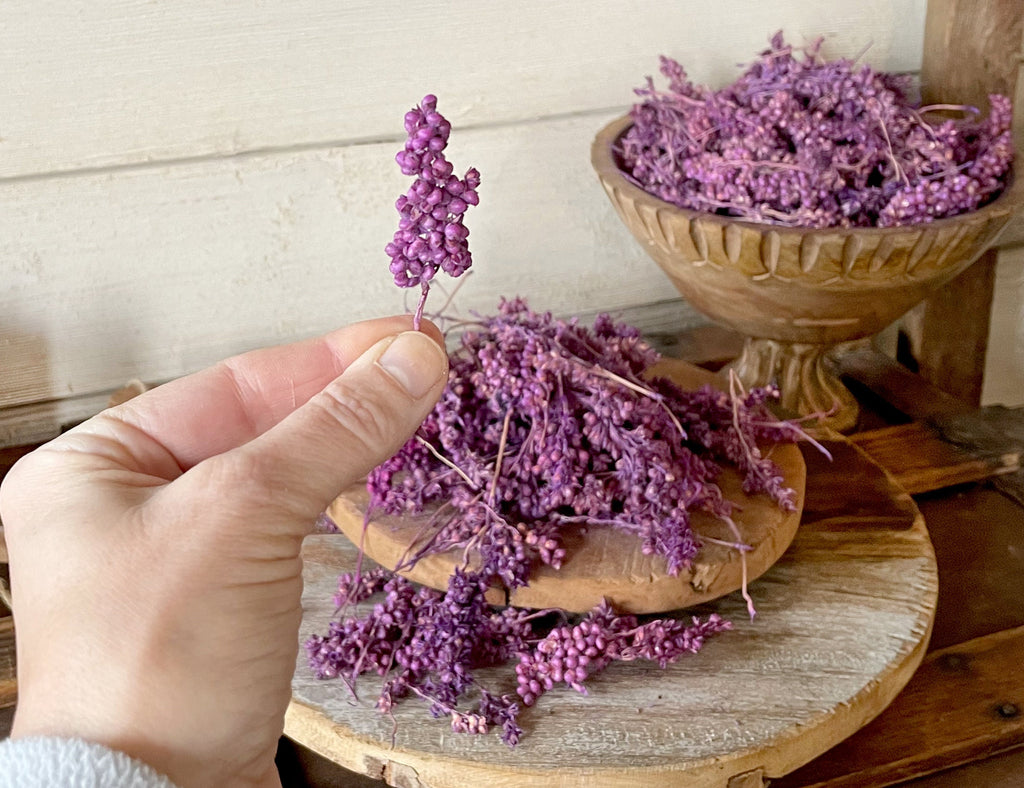 Hand holding a small bundle of purple dried flowers with a wooden bowl filled with more flowers in the background.