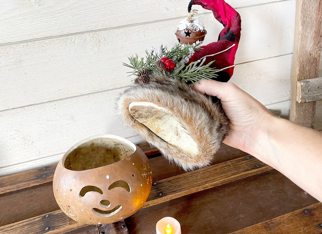 Hand holding a fur-trimmed hat with a wooden lantern and candle on a wooden surface.