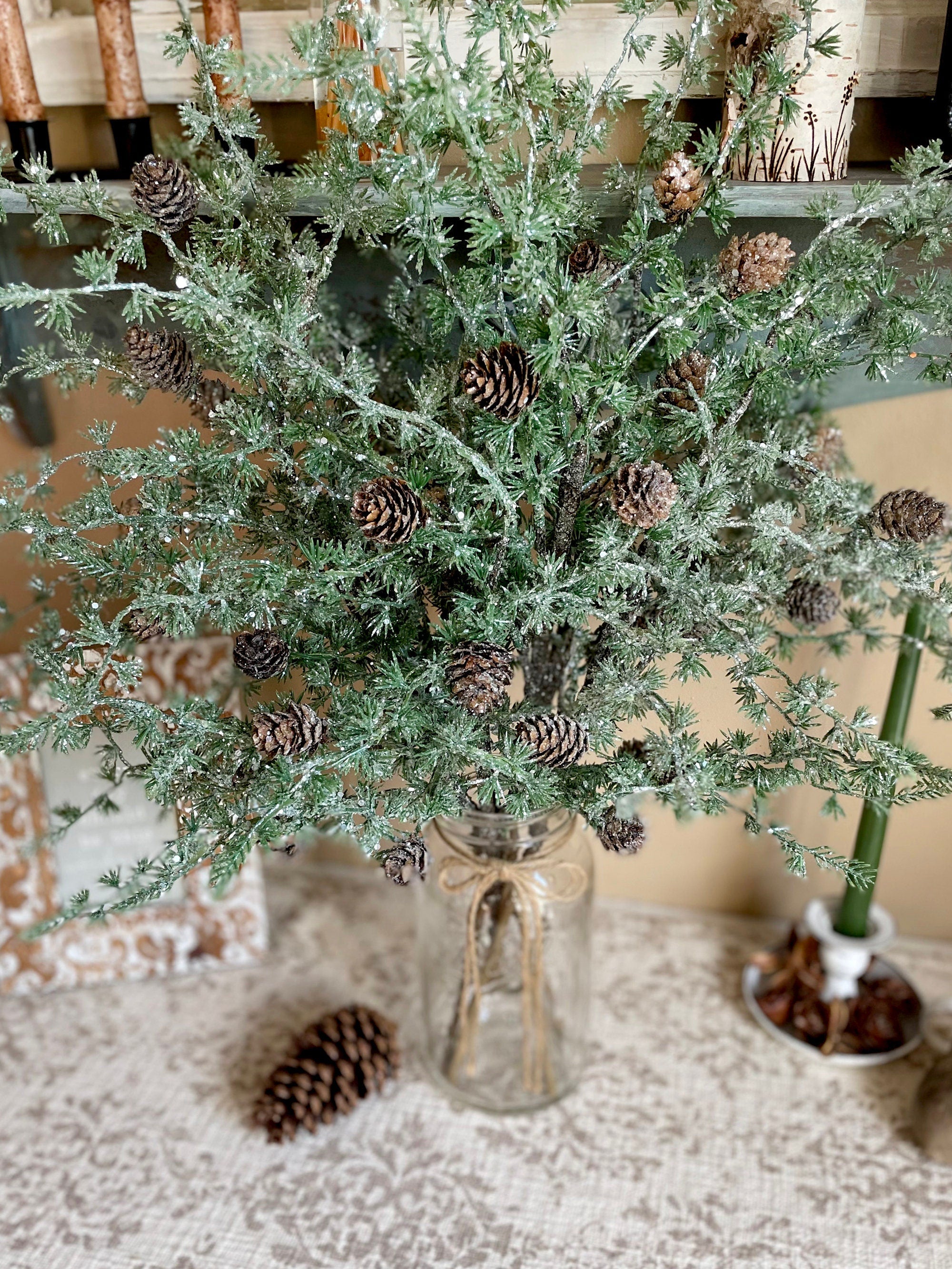 Decorative arrangement of greenery with pinecones in a glass vase on a textured surface.