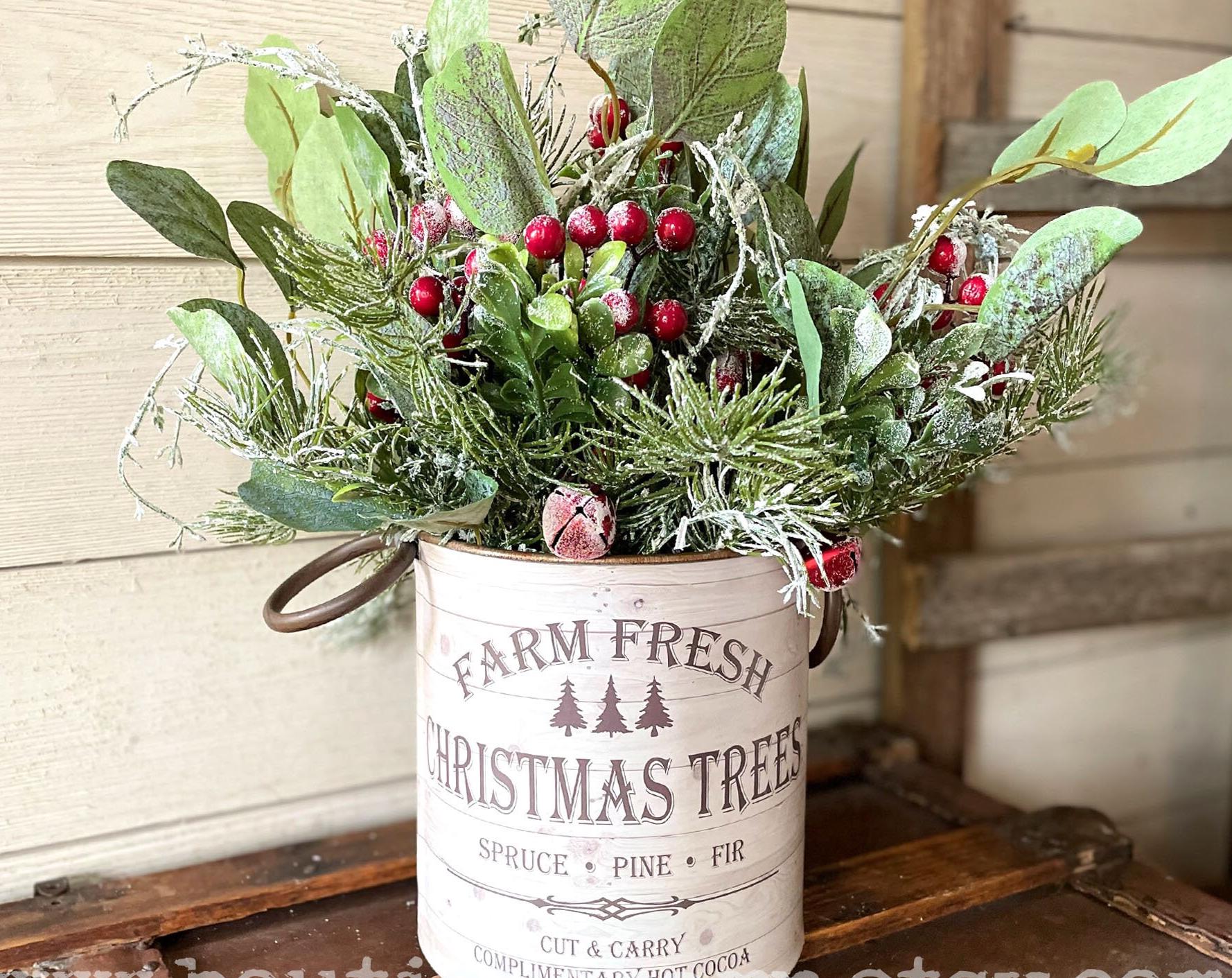 Decorative arrangement of greenery and red berries in a 'Farm Fresh Christmas Trees' container on a wooden surface.