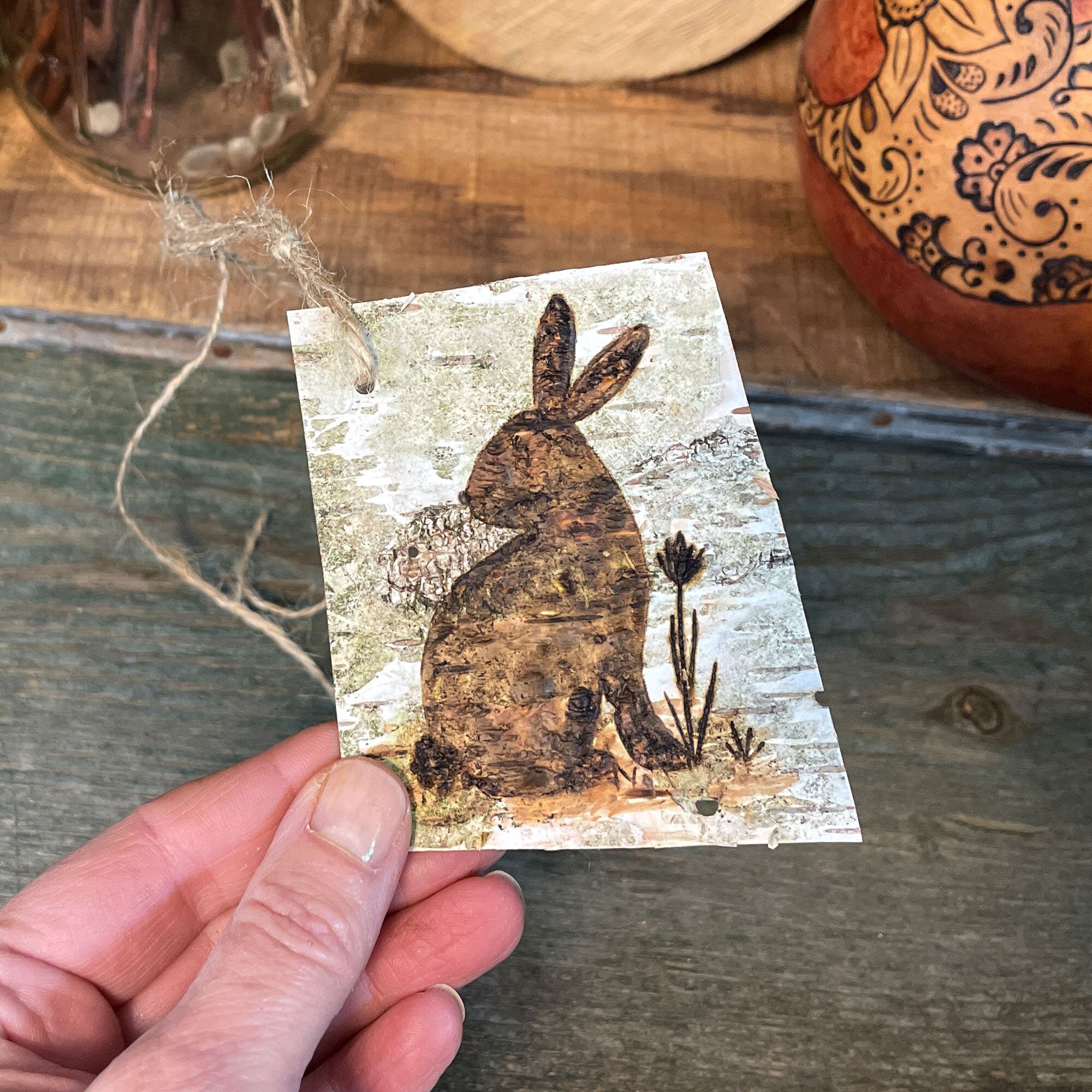 Hand holding a birch bark tag with a rabbit design on a wooden surface