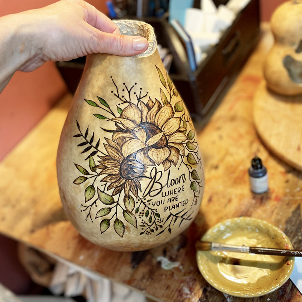 Hand holding a gourd with floral design and text on a wooden surface.