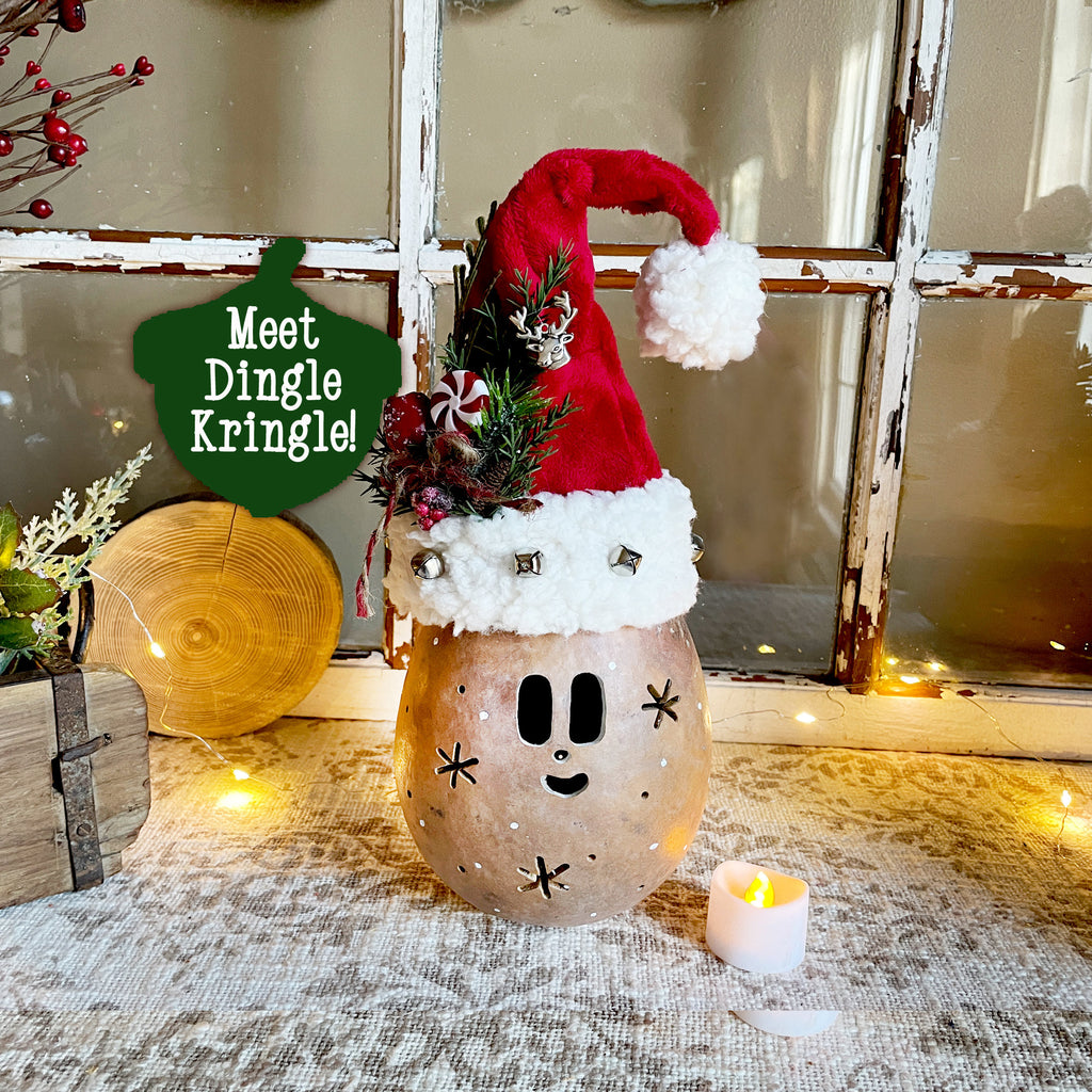 Decorative lantern with a Santa hat and face, labeled 'Meet Dingle Kringle', on a rustic surface with a candle and window in the background.