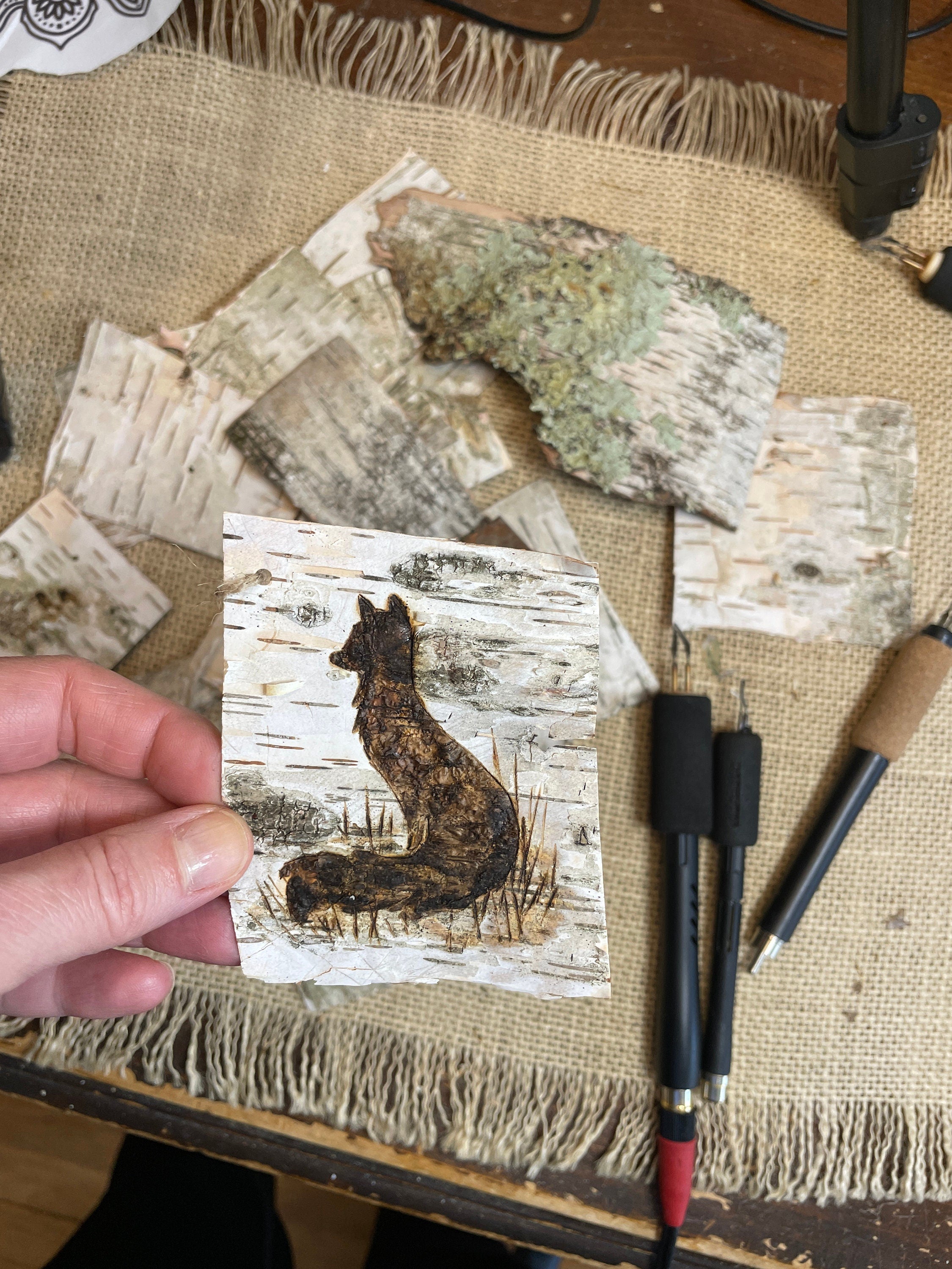 Hand holding a piece of paper with a fox illustration on a textured surface with art supplies.