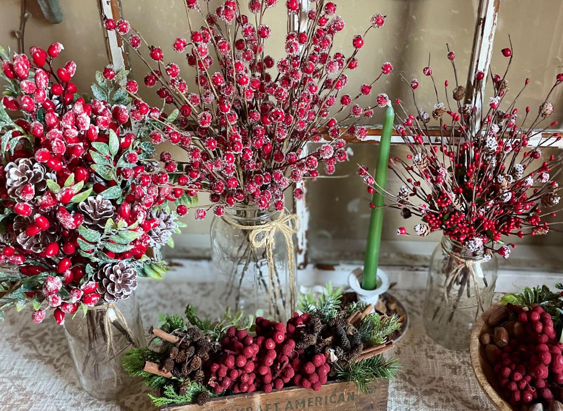 Decorative arrangement with red berries, pinecones, and a green candle in a jar on a textured surface.