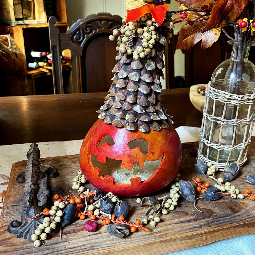 Decorative pumpkin with carved face, pine cone tree, and small figures on a wooden surface.