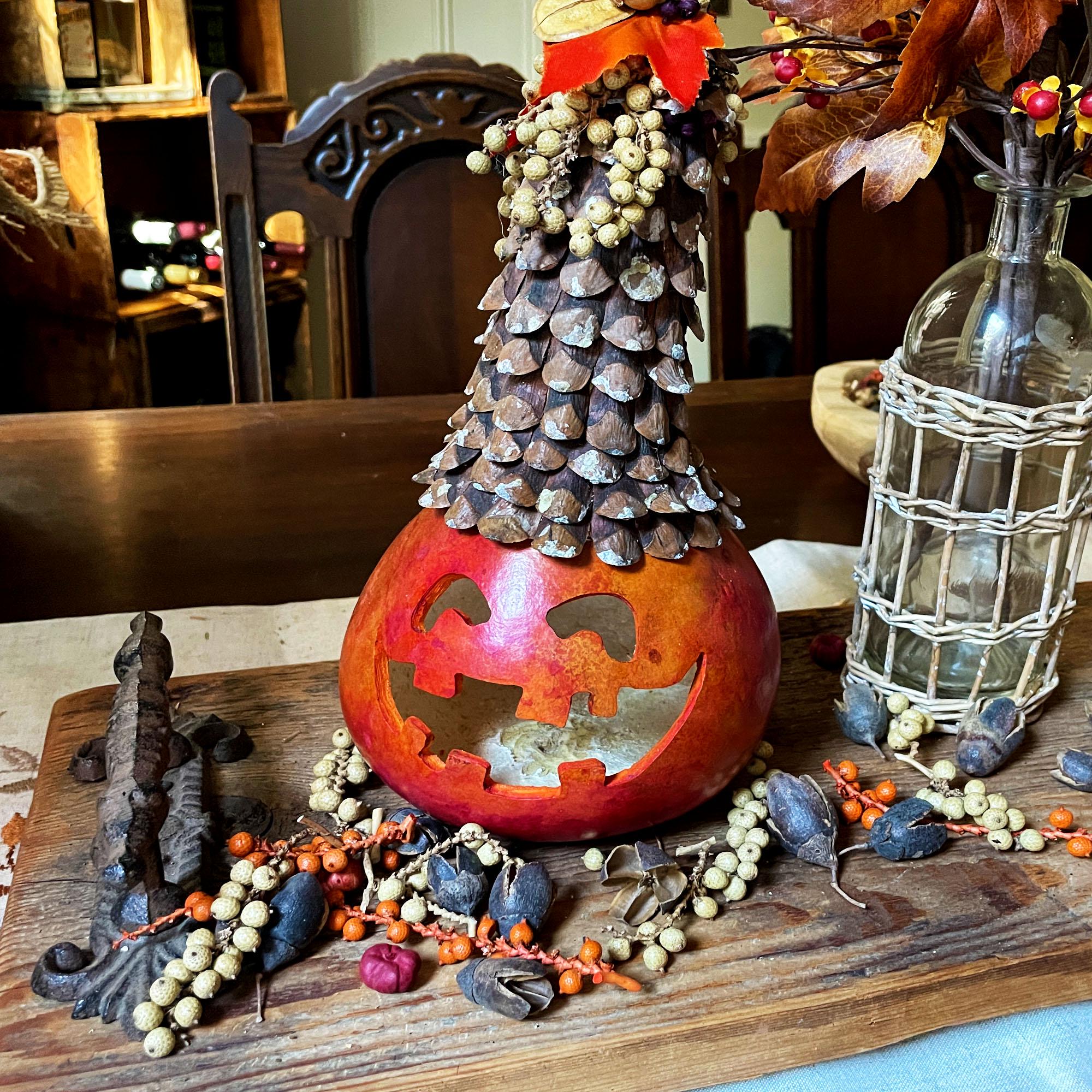 Decorative pumpkin with carved face, pine cone tree, and small figures on a wooden surface.