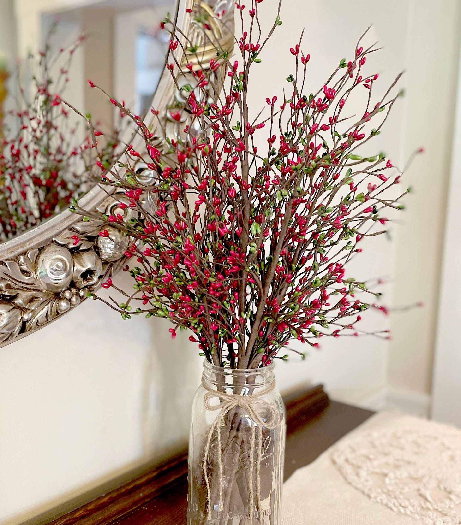 Decorative arrangement of red and green branches in a clear glass vase with a mirror in the background.