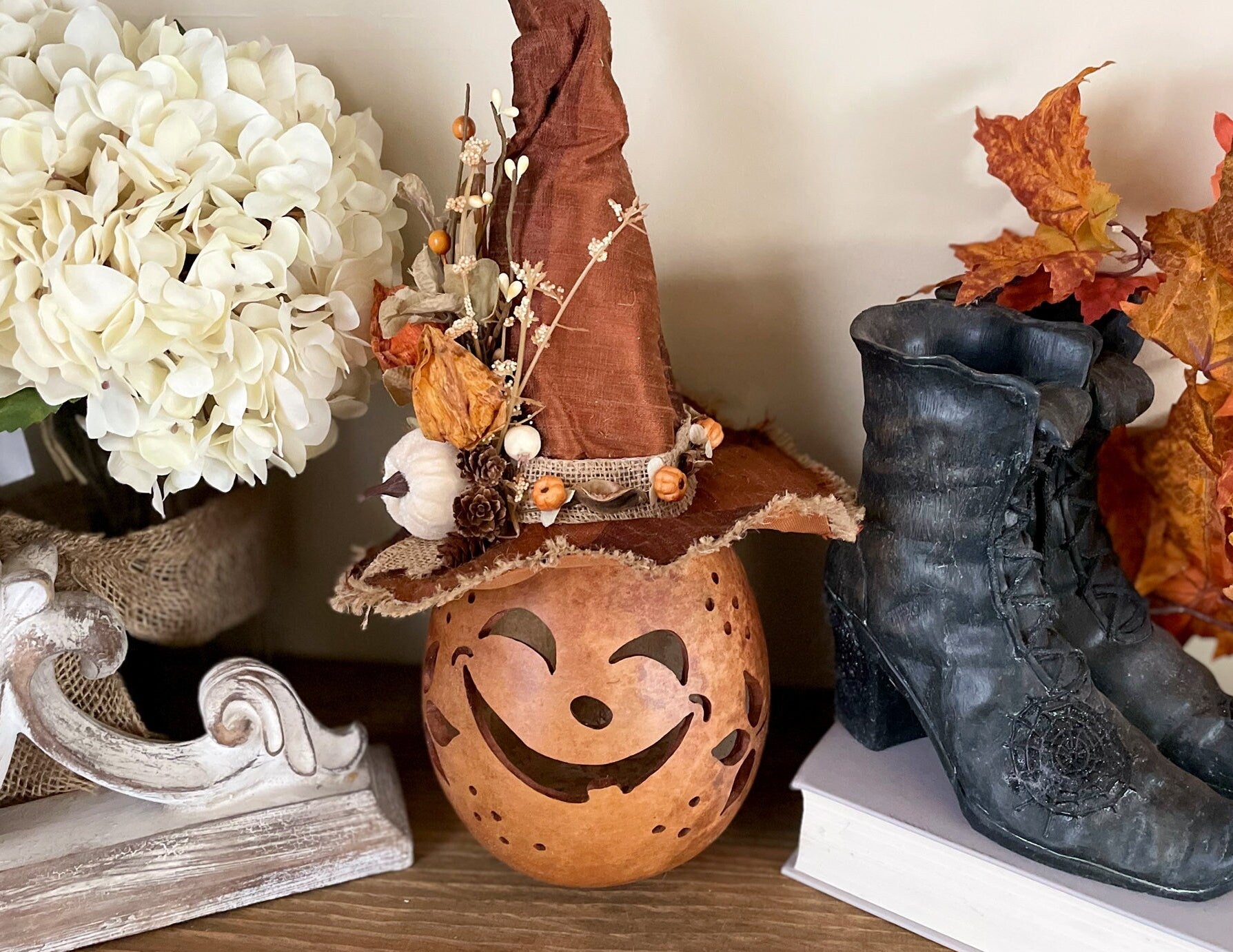 Decorative items including a pumpkin lantern, witch hat, and boots on a shelf with flowers and leaves.
