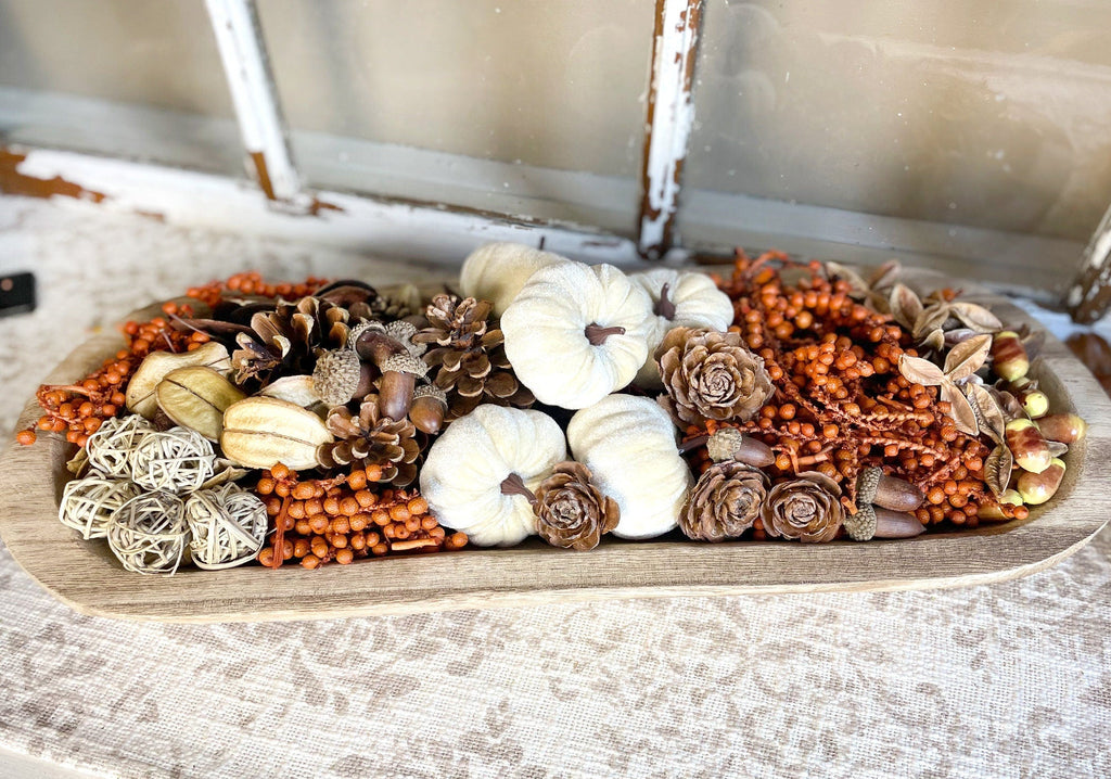 Decorative wood dough bowl arrangement of pumpkins, pinecones, and flowers on a wooden board.