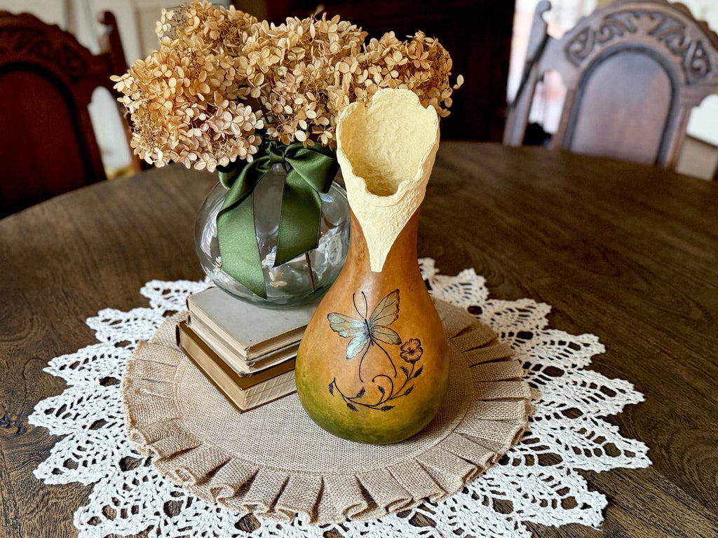 Decorative gourd with a dragonfly design on a table with a vase of flowers and books.