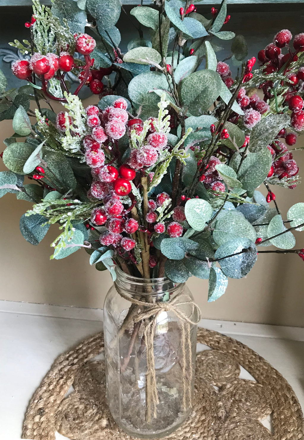 Decorative arrangement of greenery and red berries in a glass jar on a woven mat.