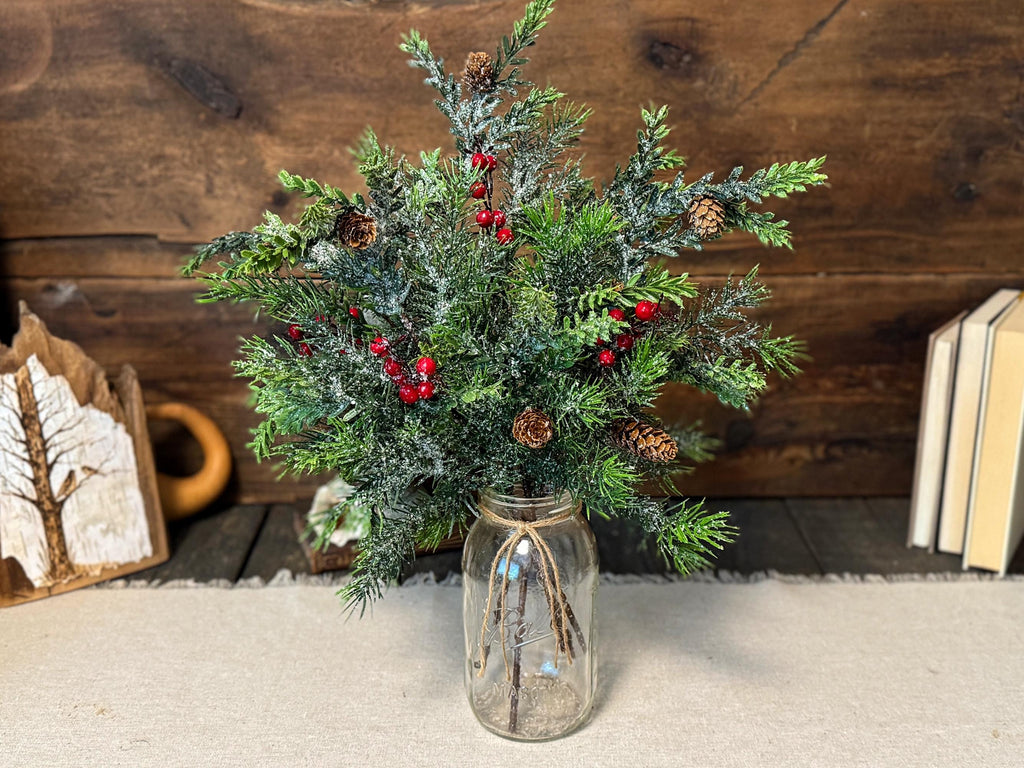 Decorative arrangement of greenery and red berries in a glass jar on a wooden surface.