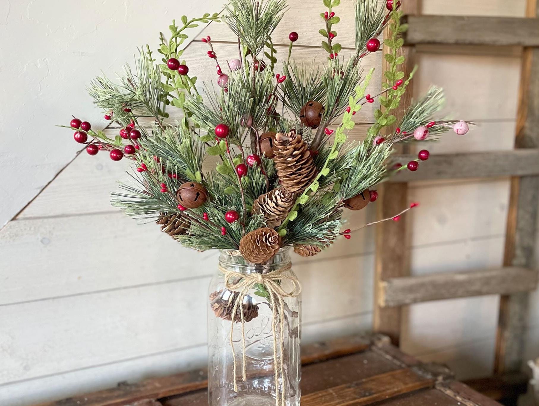 Decorative arrangement of greenery, red berries, and pinecones in a glass jar on a wooden surface.