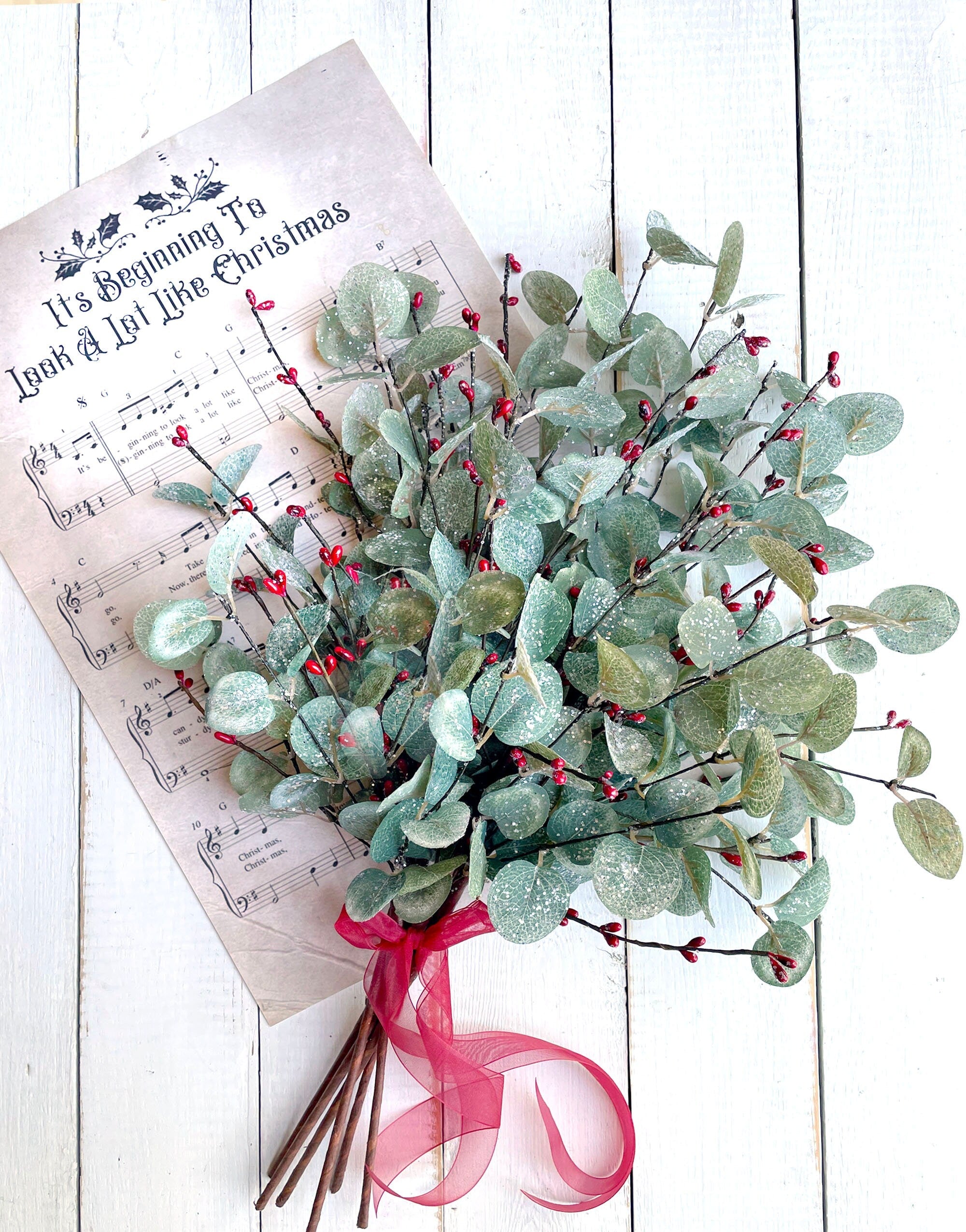 Bouquet of greenery with red berries on a sheet of music with text, on a white wooden surface.