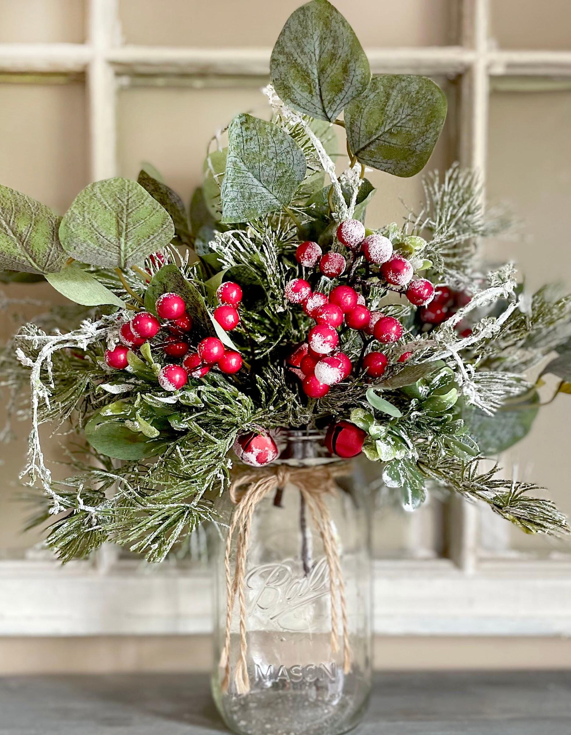 Decorative arrangement of greenery and red berries in a clear glass jar on a neutral background
