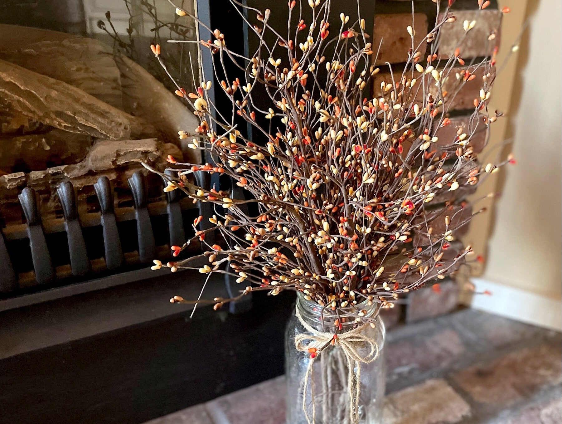 Decorative arrangement of branches with small red berries in a clear glass vase in front of a fireplace.