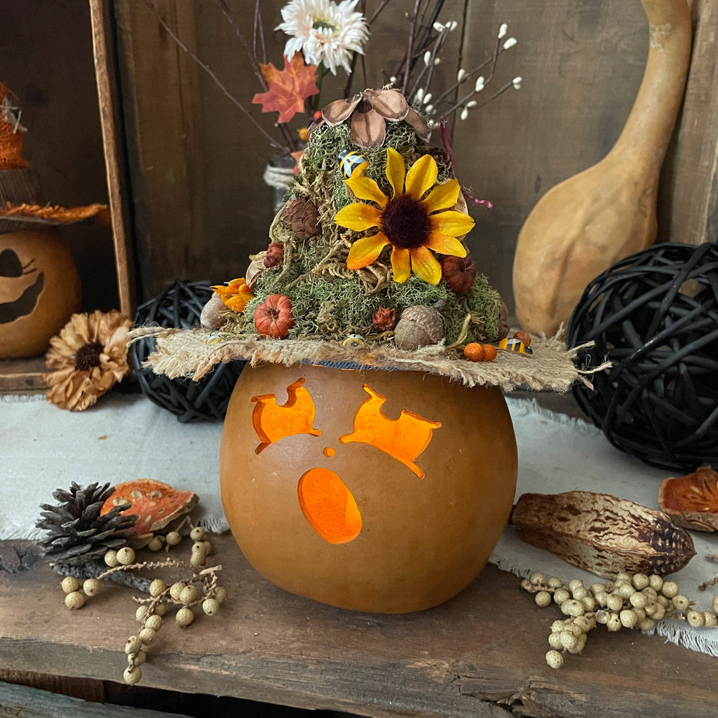 Decorative pumpkin with a carved face, sunflower, and fall-themed arrangement on a wooden surface.