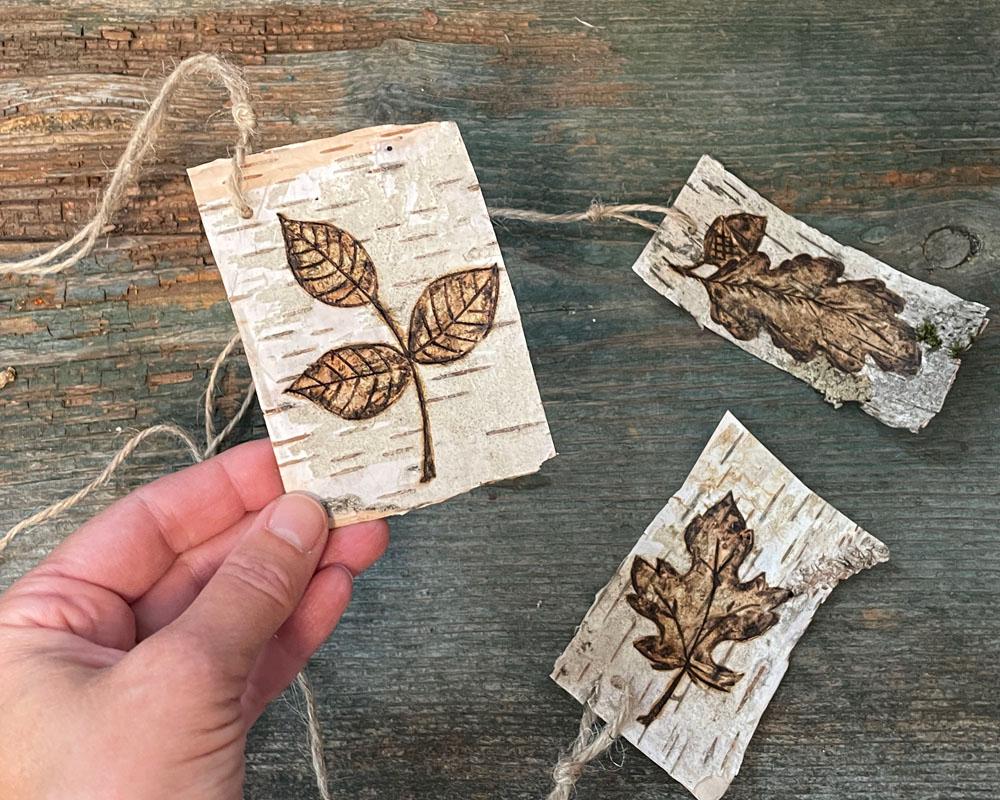 Hand holding a hand drawn leaf wood tag with two additional tags on a wooden surface.