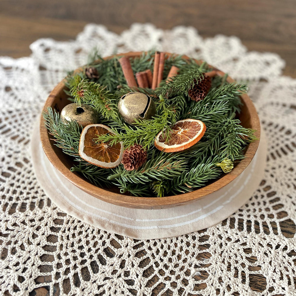 Wooden bowl filled with greenery, dried oranges, and cinnamon sticks on a lace tablecloth.