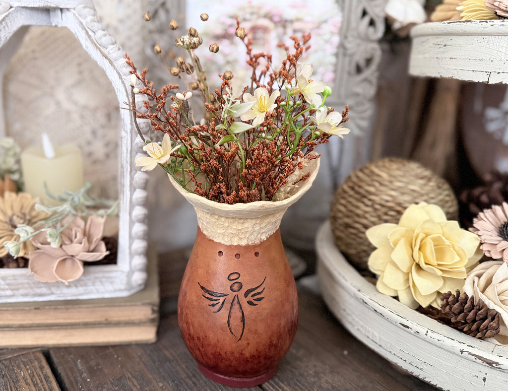 Decorative gourd vase with a dried floral arrangement. A angel is drawn on the front of the gourd. The top two inches of the rim is a beige color and the bottom is a cherry brown. 
