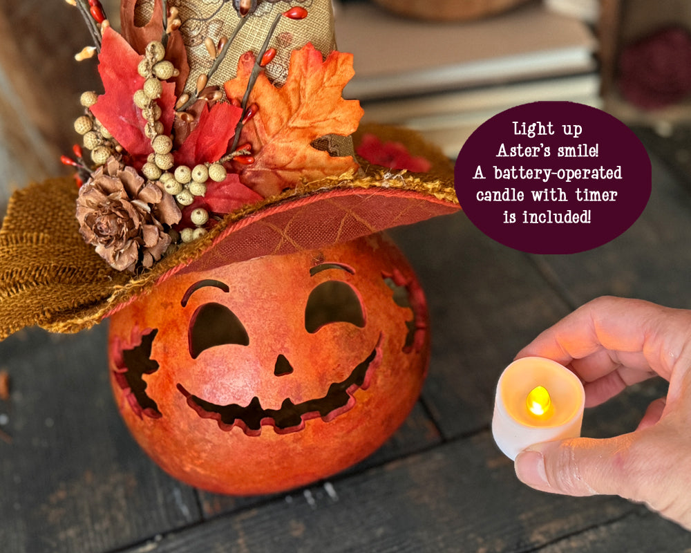 Decorative pumpkin with a smiling face, autumn leaves, and pinecones, with a hand holding a battery-operated candle.