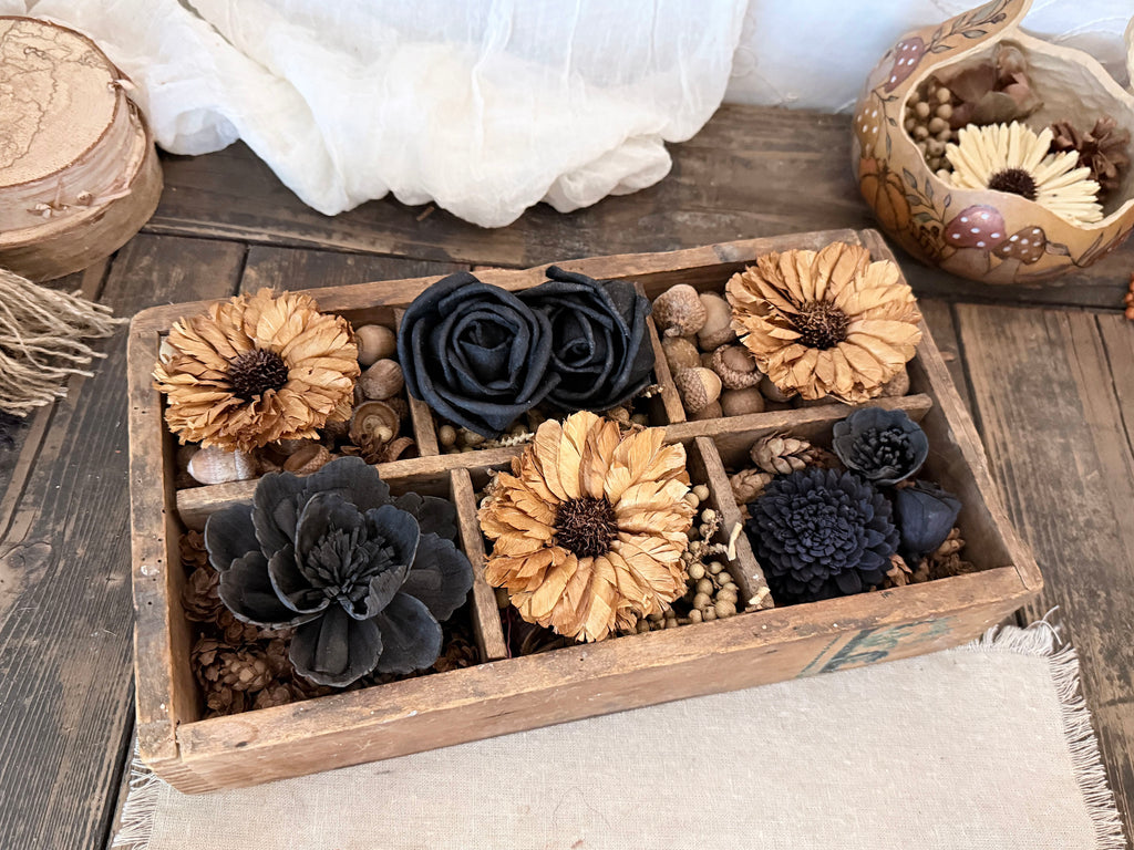 A rustic wooden box filled with dried flowers and botanical pieces in black and tan tones, styled on a wooden surface with fabric and decorative props around it.