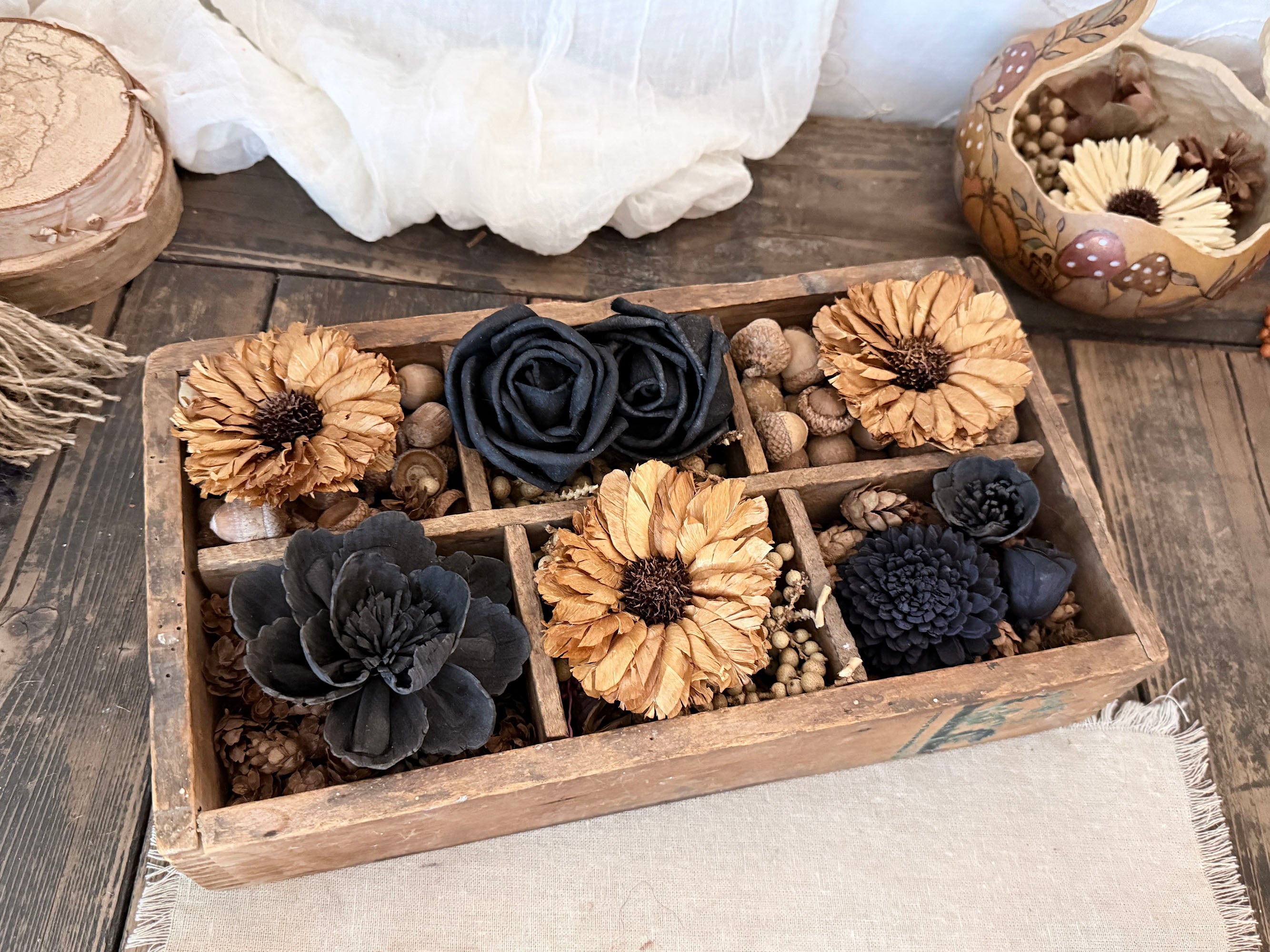A rustic wooden box filled with dried flowers and botanical pieces in black and tan tones, styled on a wooden surface with fabric and decorative props around it.