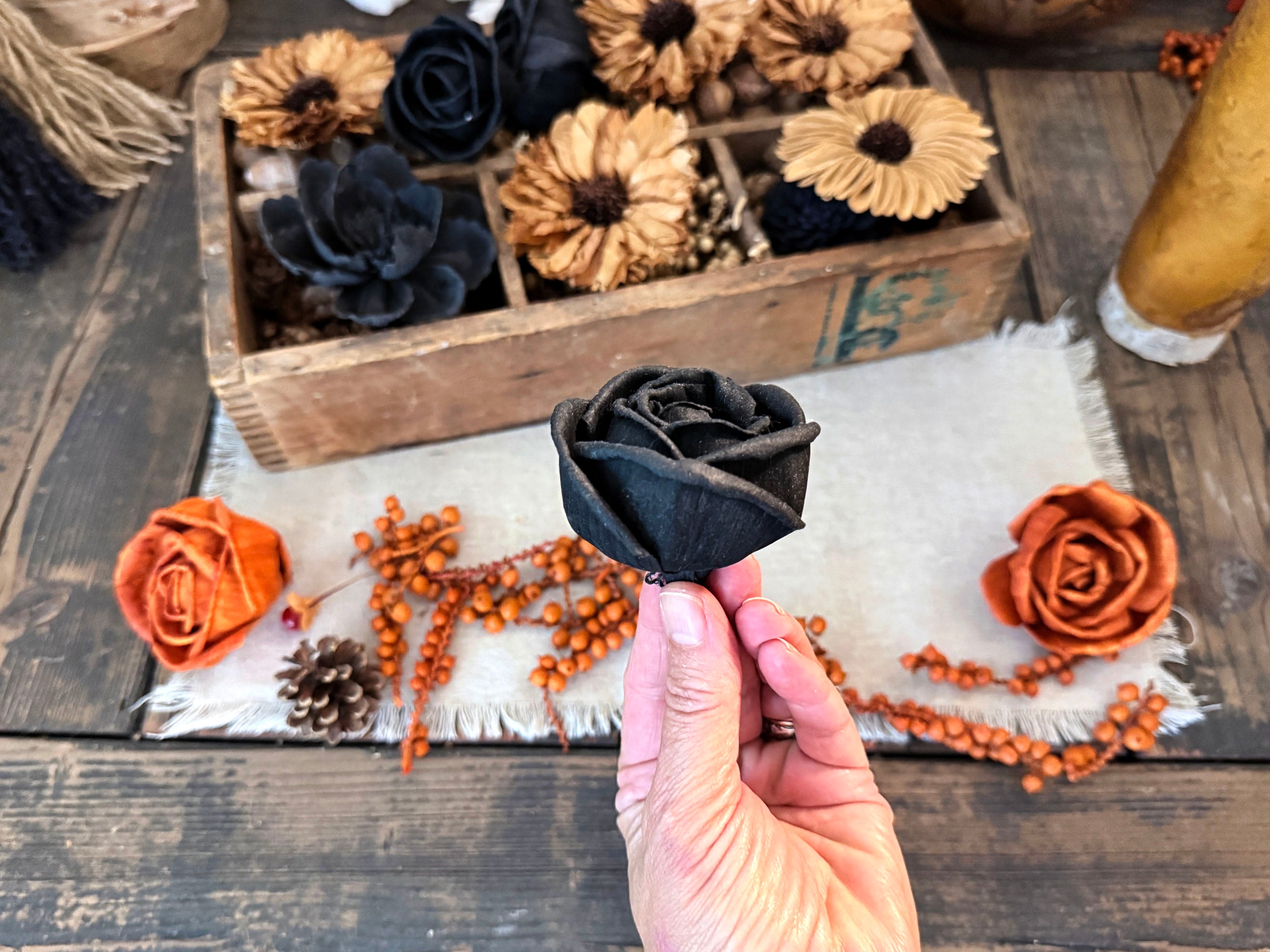 A hand holds a black wood rose blossom in the foreground, with a wooden box of dried floral decorations and orange autumn-themed decor on a rustic wooden table in the background.